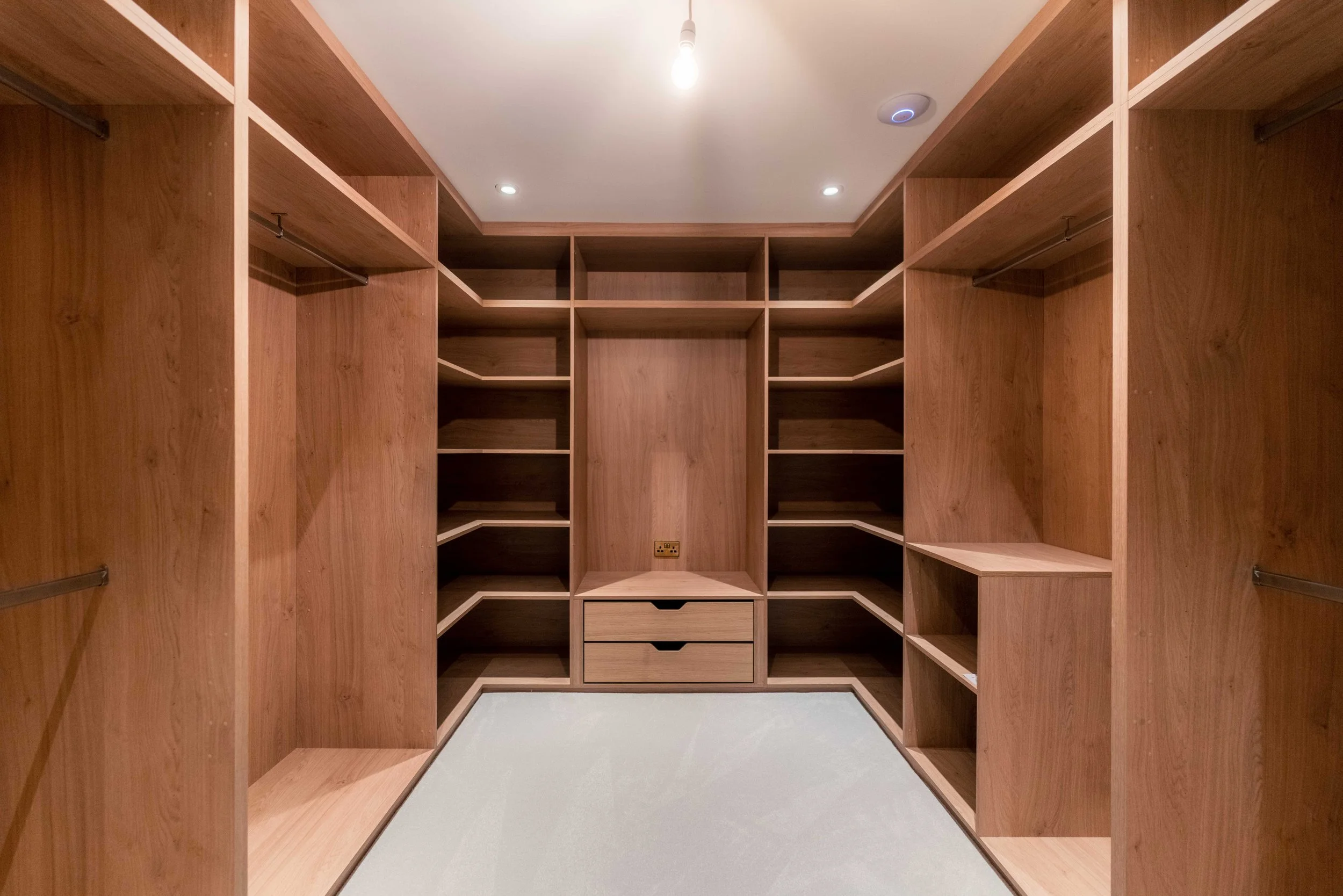 Empty walk-in closet with wooden shelves and drawers, light-colored carpet, ceiling with recessed lighting and a central smoke detector.