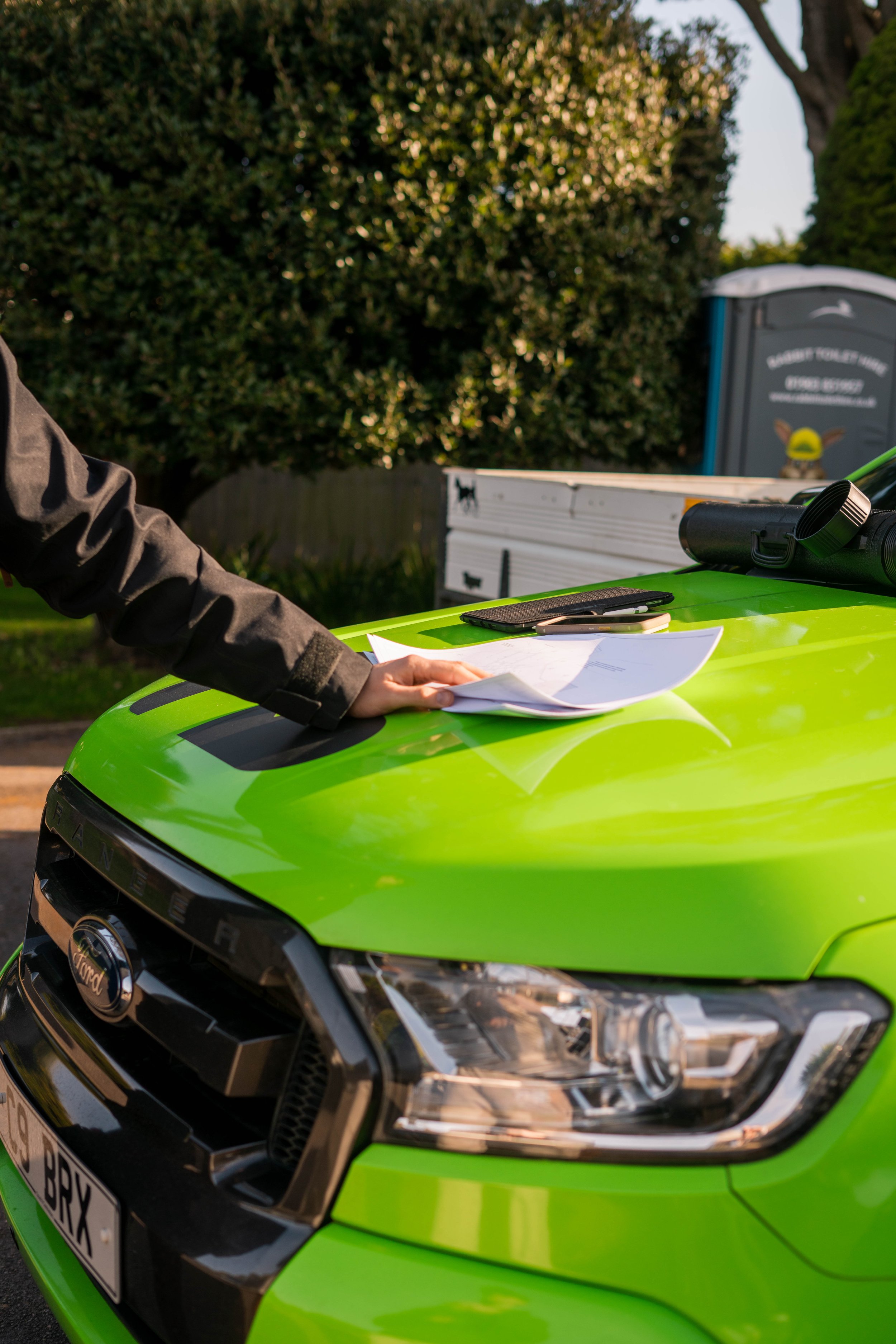 Close-up of a bright green Ford car with a person holding papers on the hood, and a smartphone placed on top. In the background, there is a portable toilet and some trees.