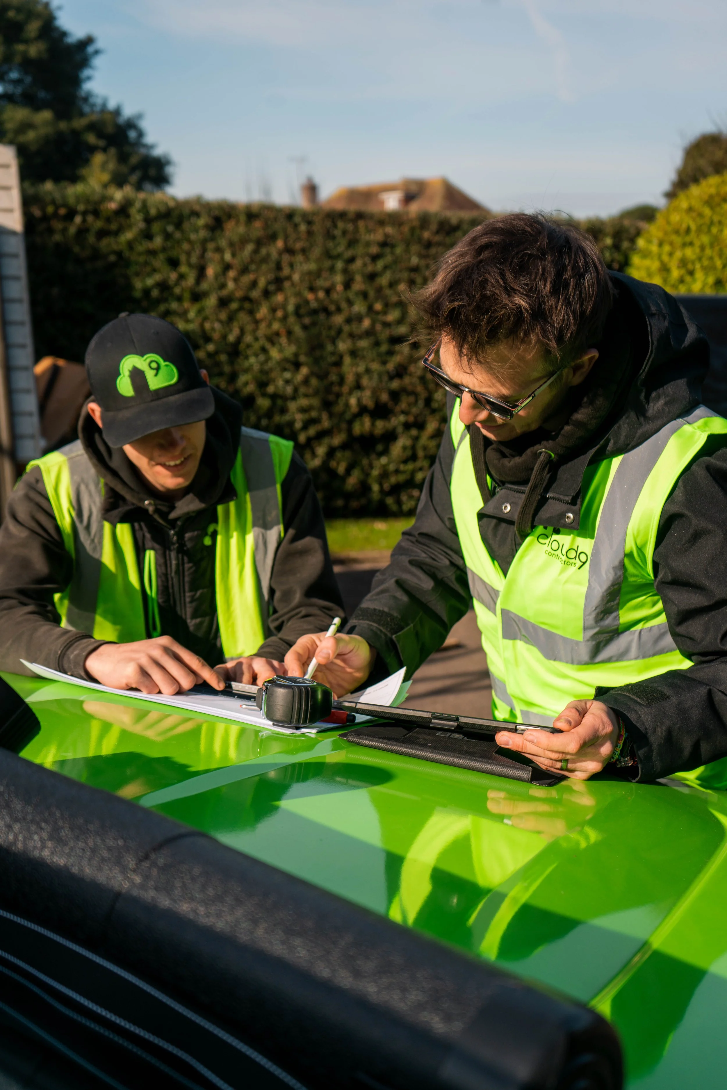 Two men wearing safety vests and black jackets are working together outside, leaning over a table with paperwork and a car key, with a hedge and houses in the background.
