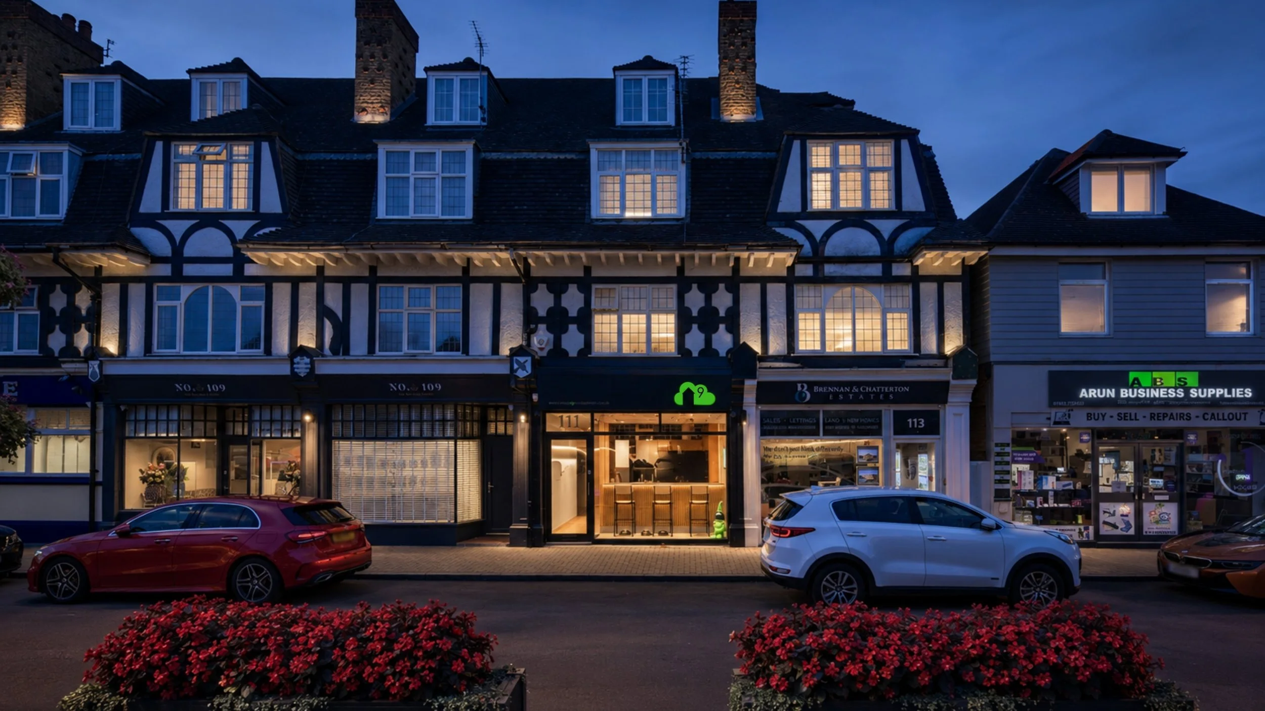 A row of storefronts on a city street at dusk with lit windows and cars parked in front, including an estate agency, a business supplies store, and a cafe or bar with warm interior lighting.