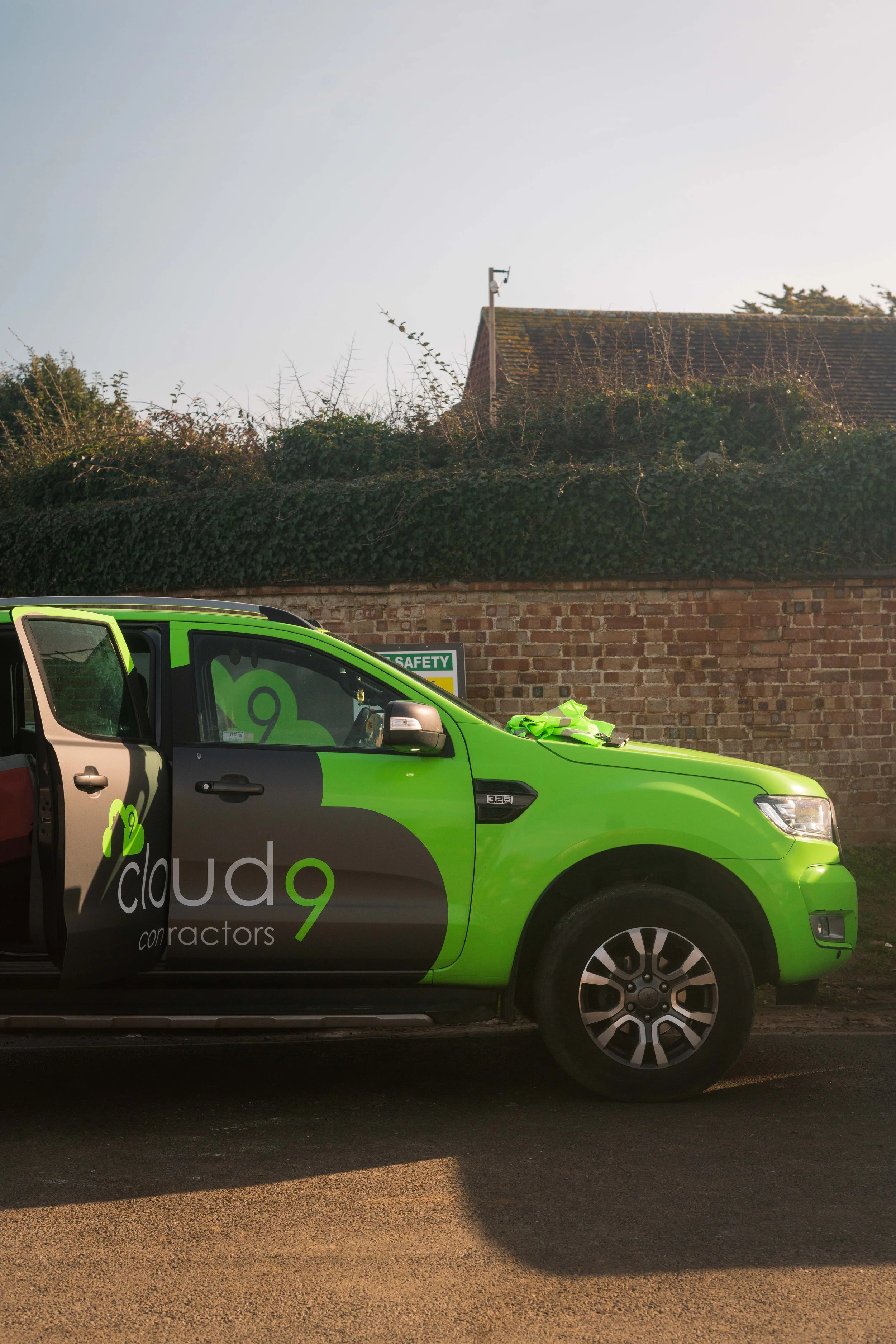 A green and black branded contractor vehicle with a logo of a cloud and the words 'cloud 9 contractors' parked on a street in front of a brick wall topped with greenery, with a house roof and a weather monitoring device visible in the background.