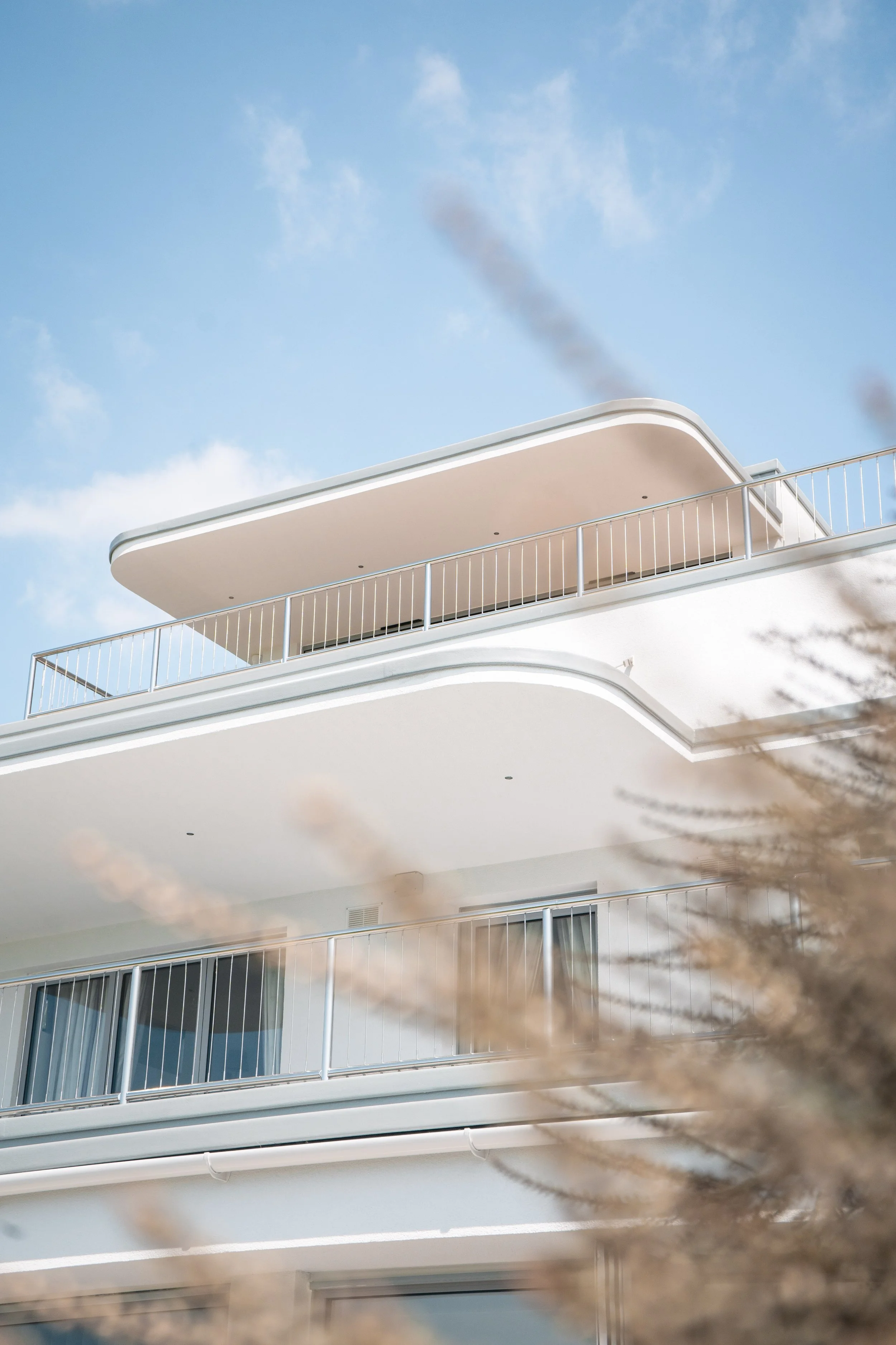 Close-up view of modern white apartment building balconies against a bright blue sky with some clouds, with blurred tree branches in the foreground.