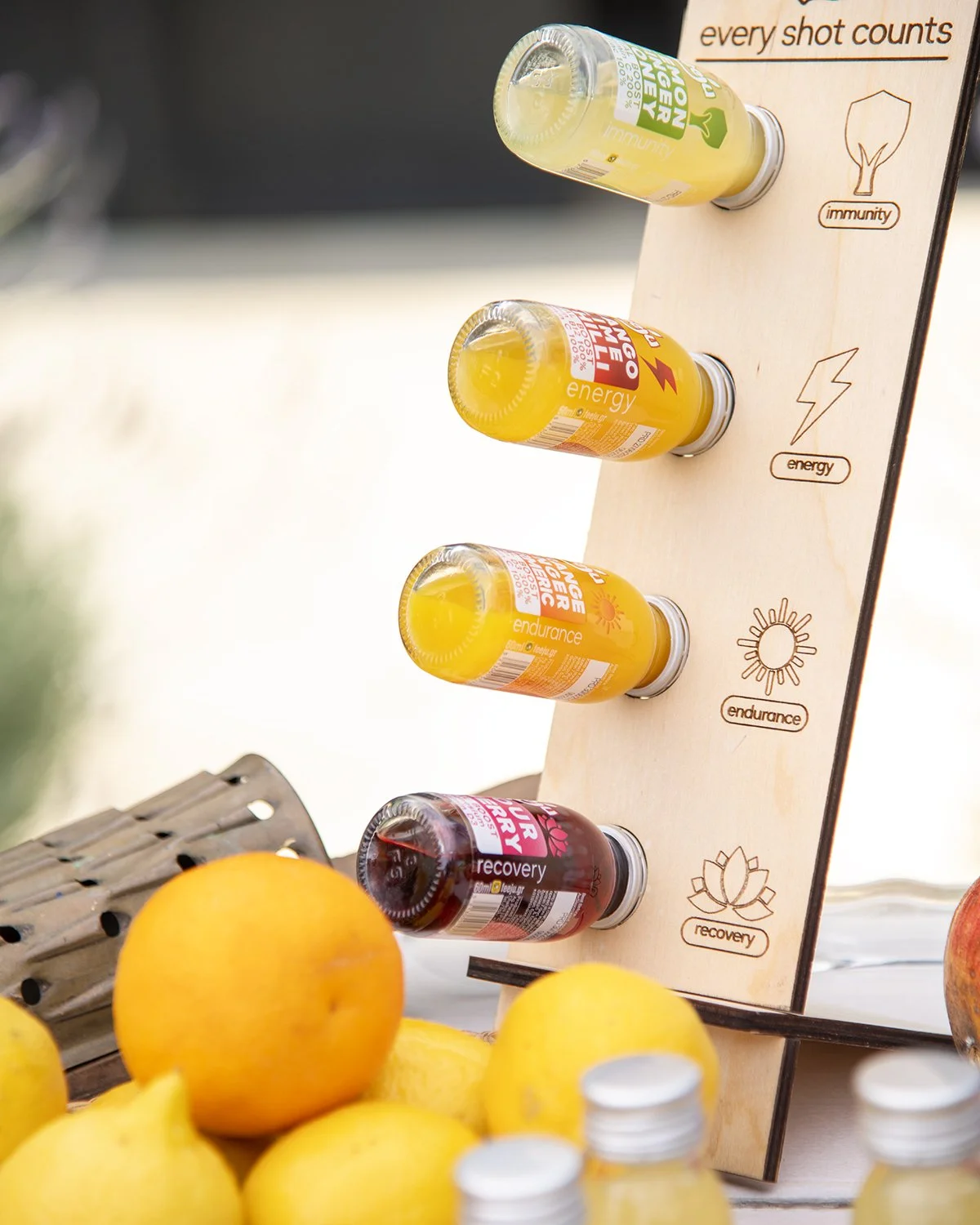 A display of various vitamin and supplement shots labeled as immunity, energy, endurance, and recovery on a wooden board, with oranges and a small wooden brush nearby.