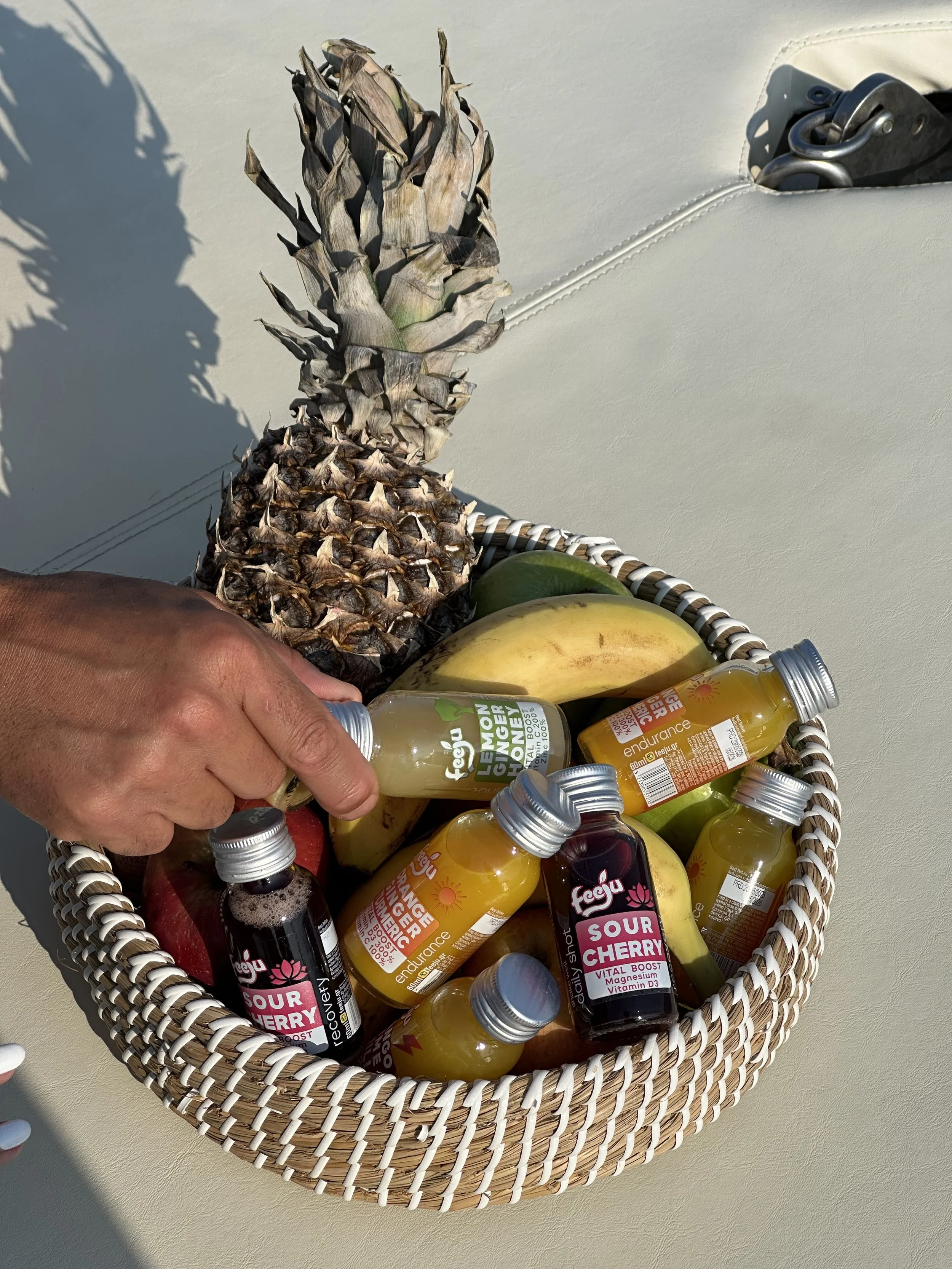A woven basket filled with bananas, pineapples, apples, and bottled beverages, seen on a beige surface outdoors with sunlight casting shadows.