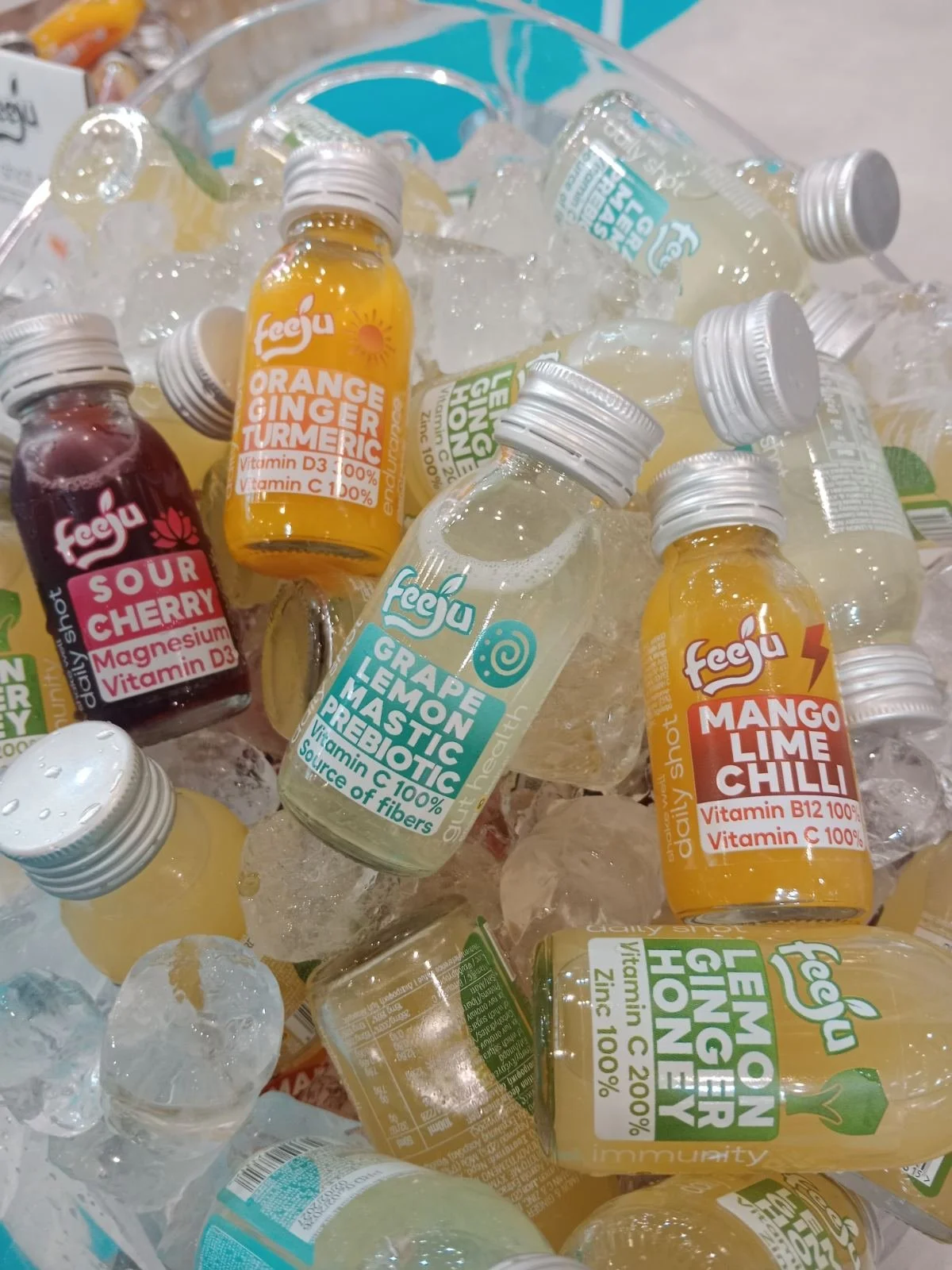 Assorted bottled herbal and fruit drinks in a container with ice, including orange ginger turmeric, sour cherry, grape lemon tonic, mango lime chili, and lemon ginger honey flavors.