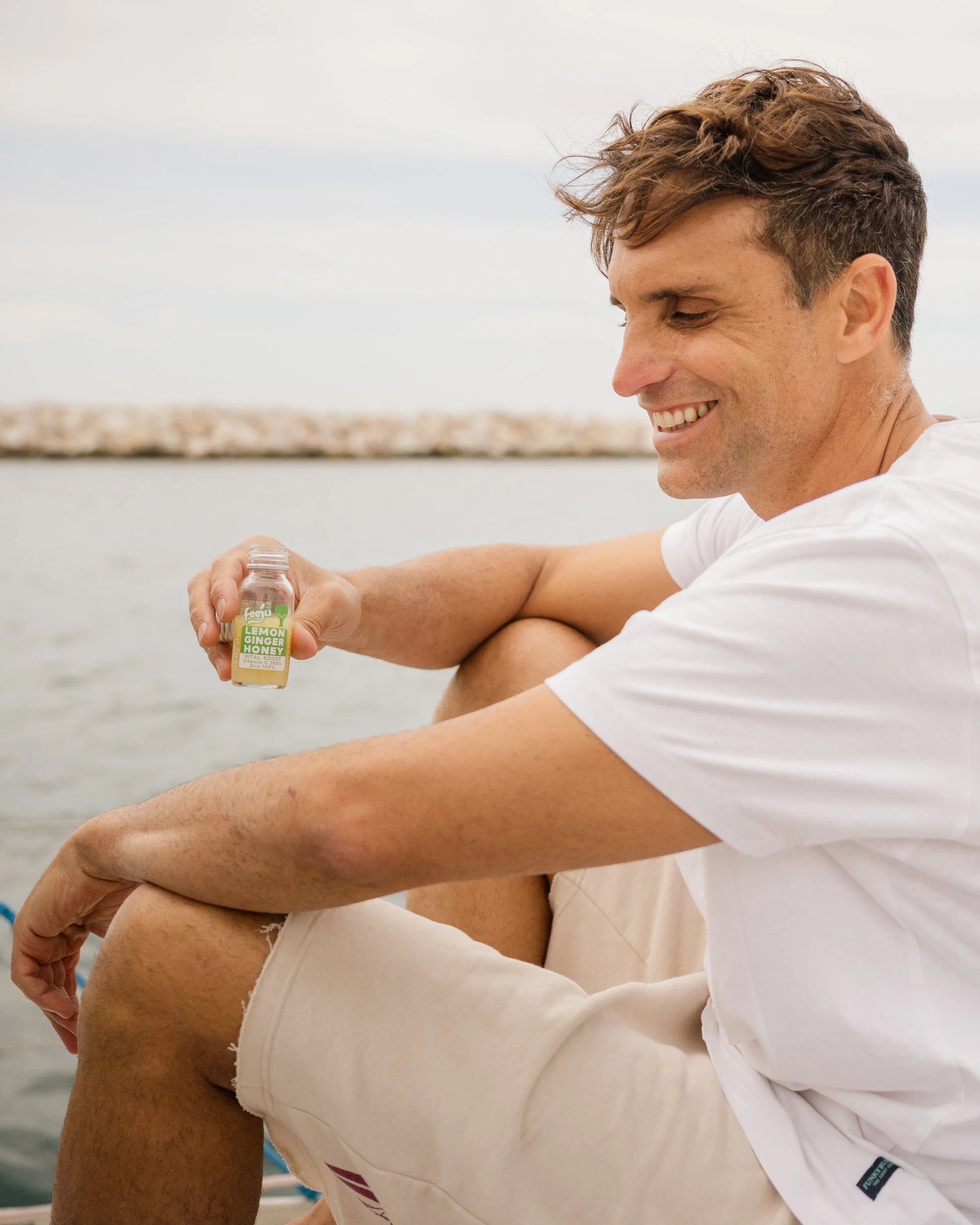 A man sitting outdoors near water, smiling, holding a lemon ginger honey drink, with a view of the water and sky in the background.