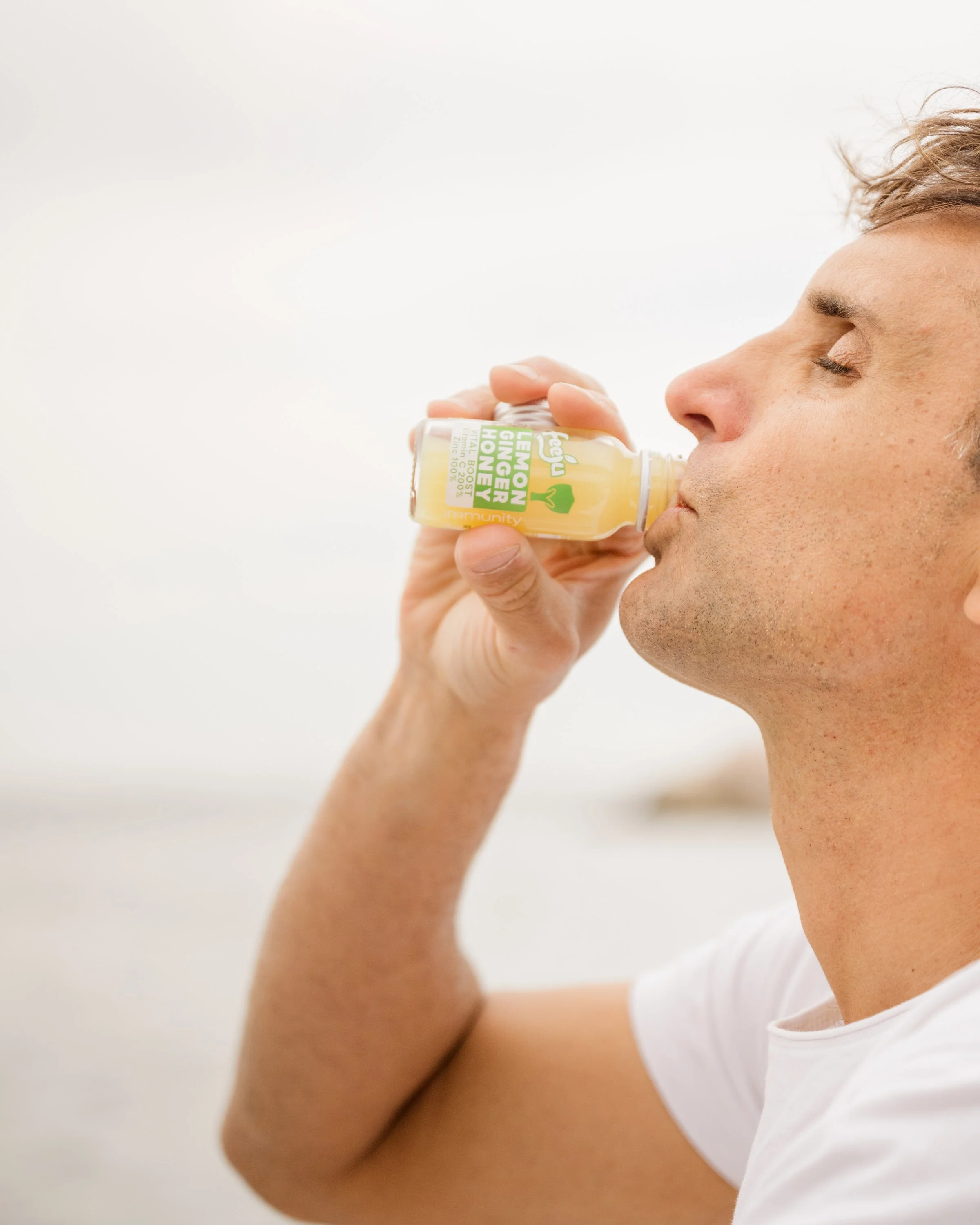 A person with light skin and short, light brown hair is seen close to the camera, with eyes closed, holding a bottle of lemon ginger honey vitamin boost close to their lips, in a bright, neutral environment.