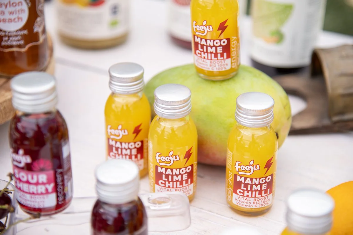 Multiple small bottles of mango lime chili juice and cherry syrup on a table with a large green fruit in the background.