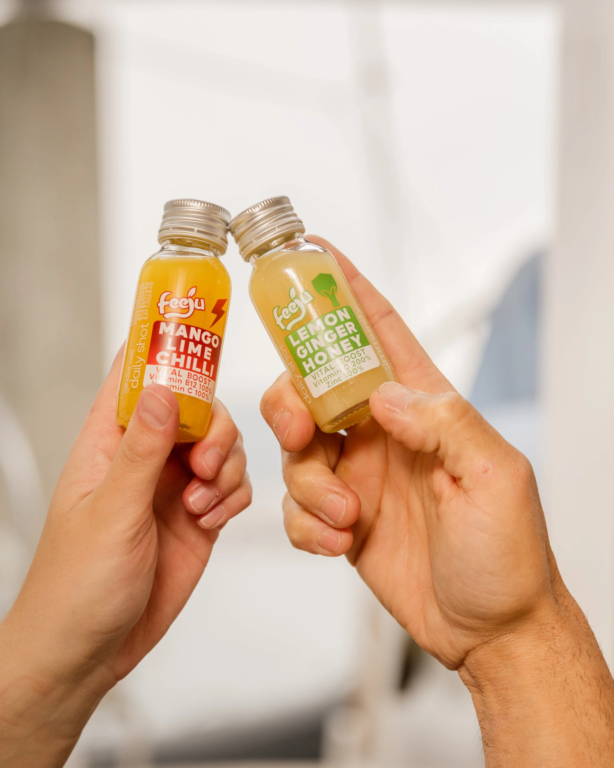 Two small bottles of health supplements are being held. The left bottle is labeled 'Faeja Mango Lime Chili', and the right bottle is labeled 'Lemon Ginger Honey'.