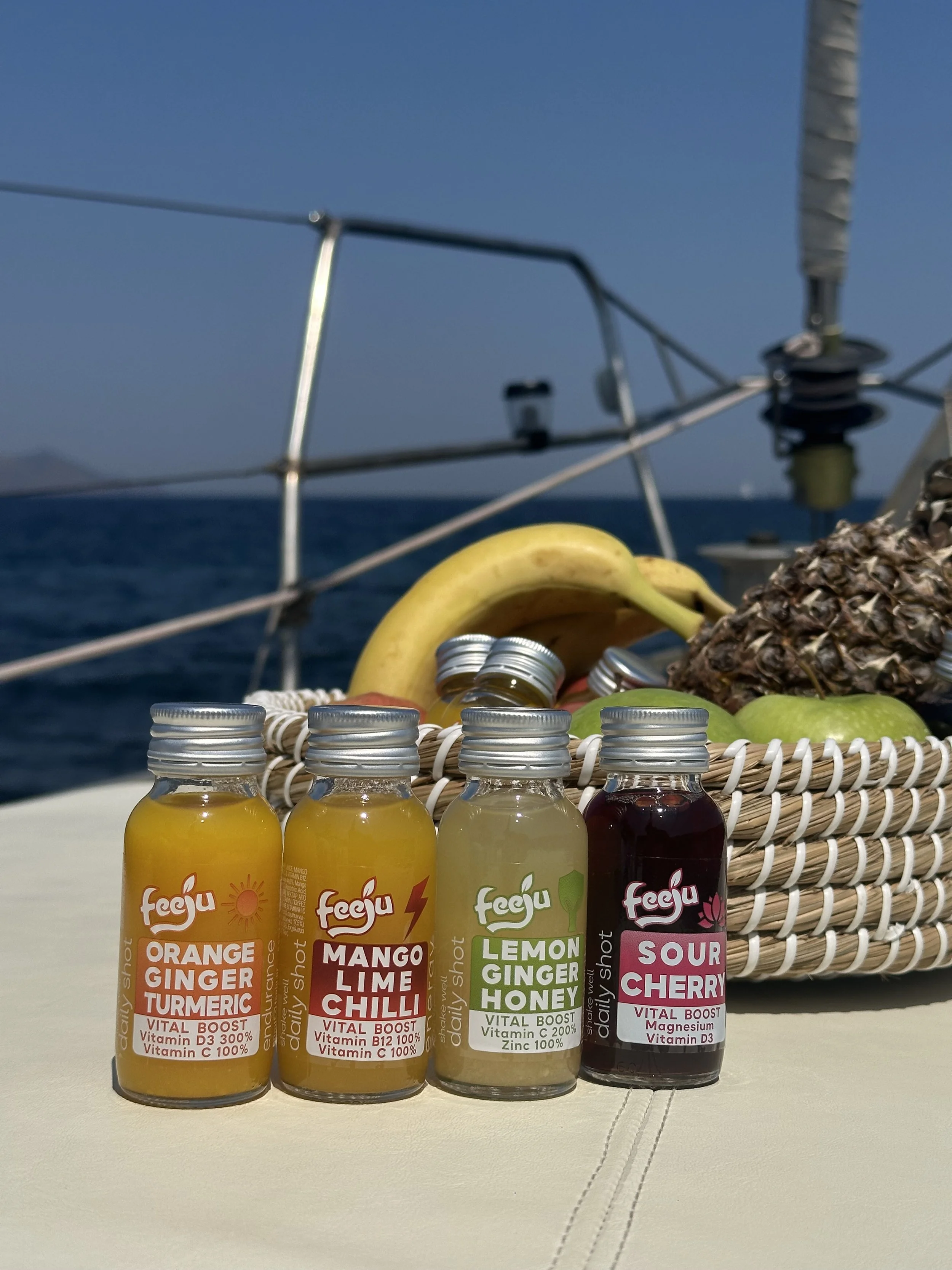 Four small bottles of Juice with flavor labels: Orange Ginger Turmeric, Mango Lime Chili, Lemon Ginger Honey, Sour Cherry, placed on a beige surface in front of a woven basket containing bananas, a pineapple, and other fruits, with a yacht's sailboat and blue sky in the background.