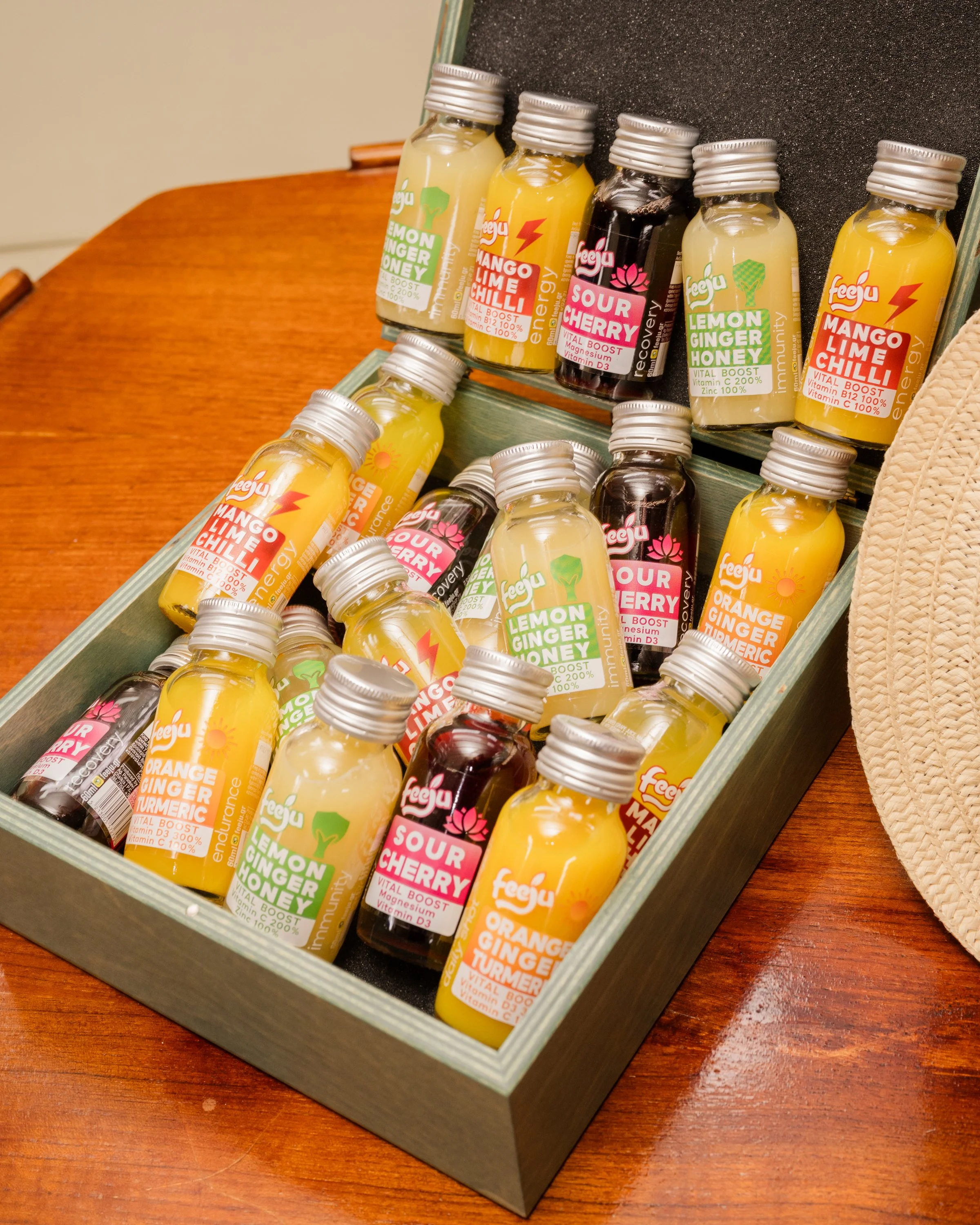 A display tray with small glass bottles of various flavors of health shots on a wooden table. Flavors include lemon ginger honey, mango lime chili, sour cherry, and orange ginger turmeric, with colorful labels and metal caps.