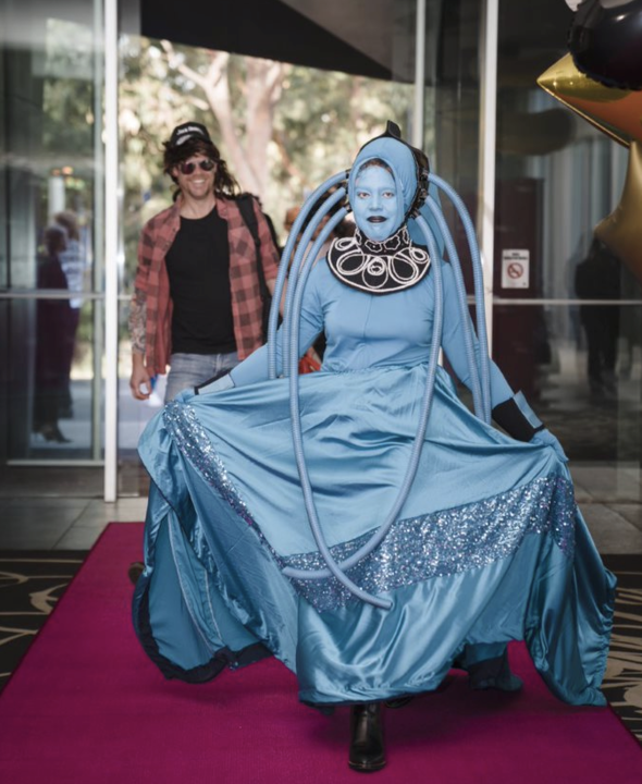 Nadia dressed as a Diva Plavalaguna costume she made, wearing a large blue gown with sparkles and a black necklace, standing on a pink carpet inside a building at the entrance of SEEK Christmas party in 2019.