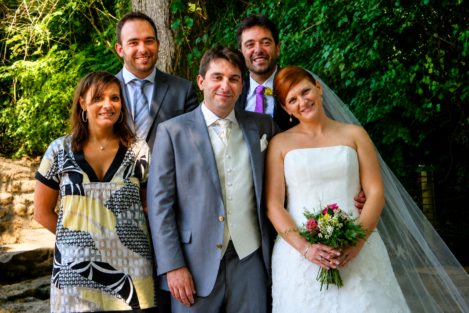 groupe de six personnes lors d'un mariage en extérieur, avec un décor verdoyant, comprenant un homme en costume gris clair, une femme en robe de mariée blanche avec un bouquet de fleurs, et quatre autres personnes, deux hommes en costumes et deux femmes