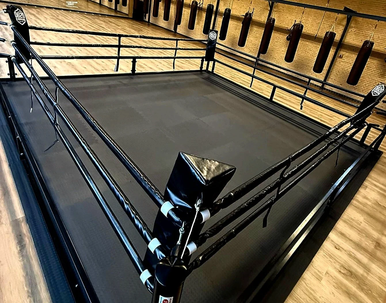 Een leeg bokszakring in een sportschool met houten vloer, omringd door trainingszolen en een muur met meerdere bokszakken.