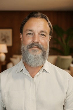 A middle-aged man with a beard and mustache, wearing a white button-up shirt, standing in a warmly lit living room with wooden decor and a large plant in the background.