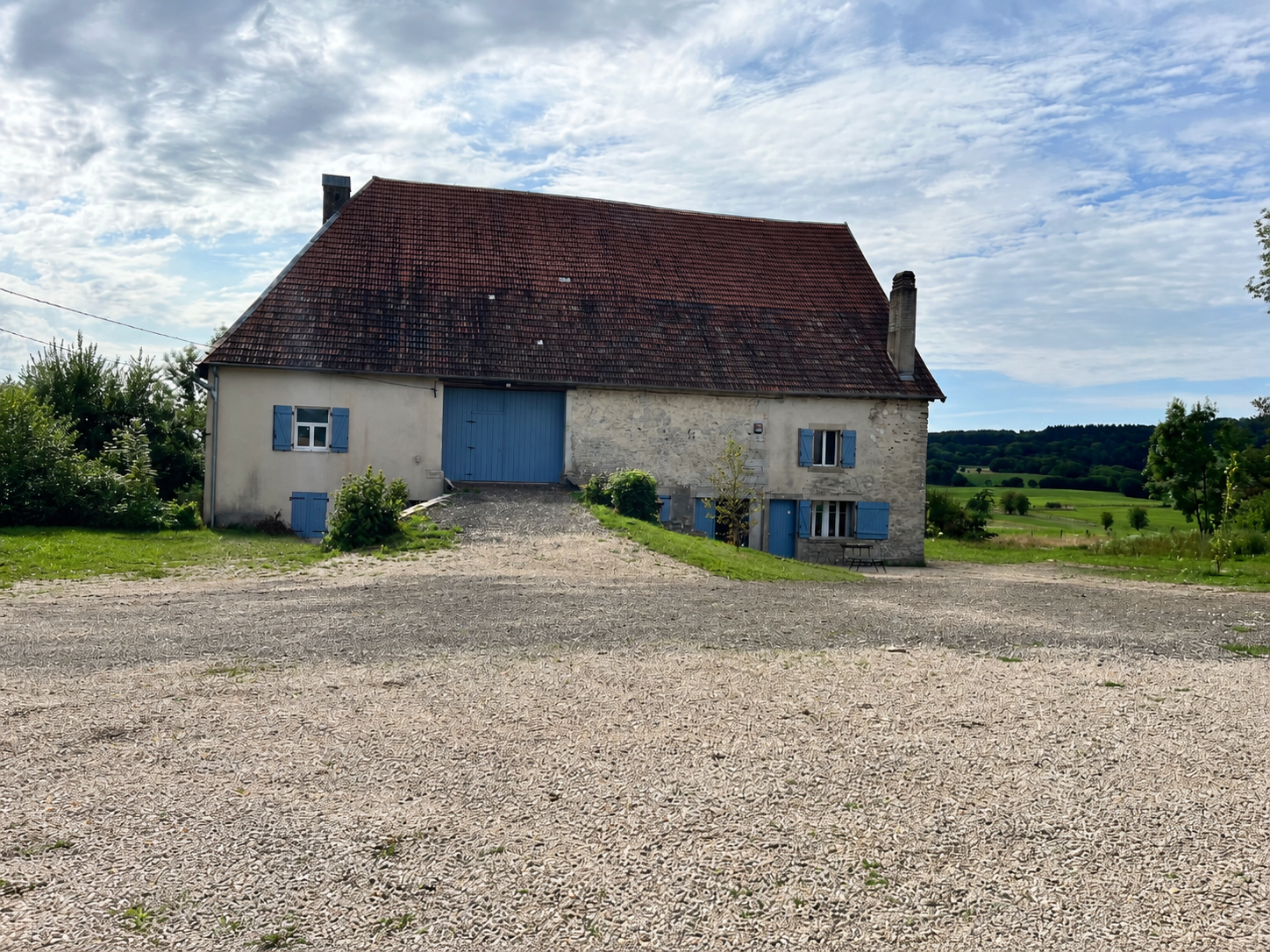 Maison en pierre avec volets bleus dans un paysage rural sous un ciel partiellement nuageux.