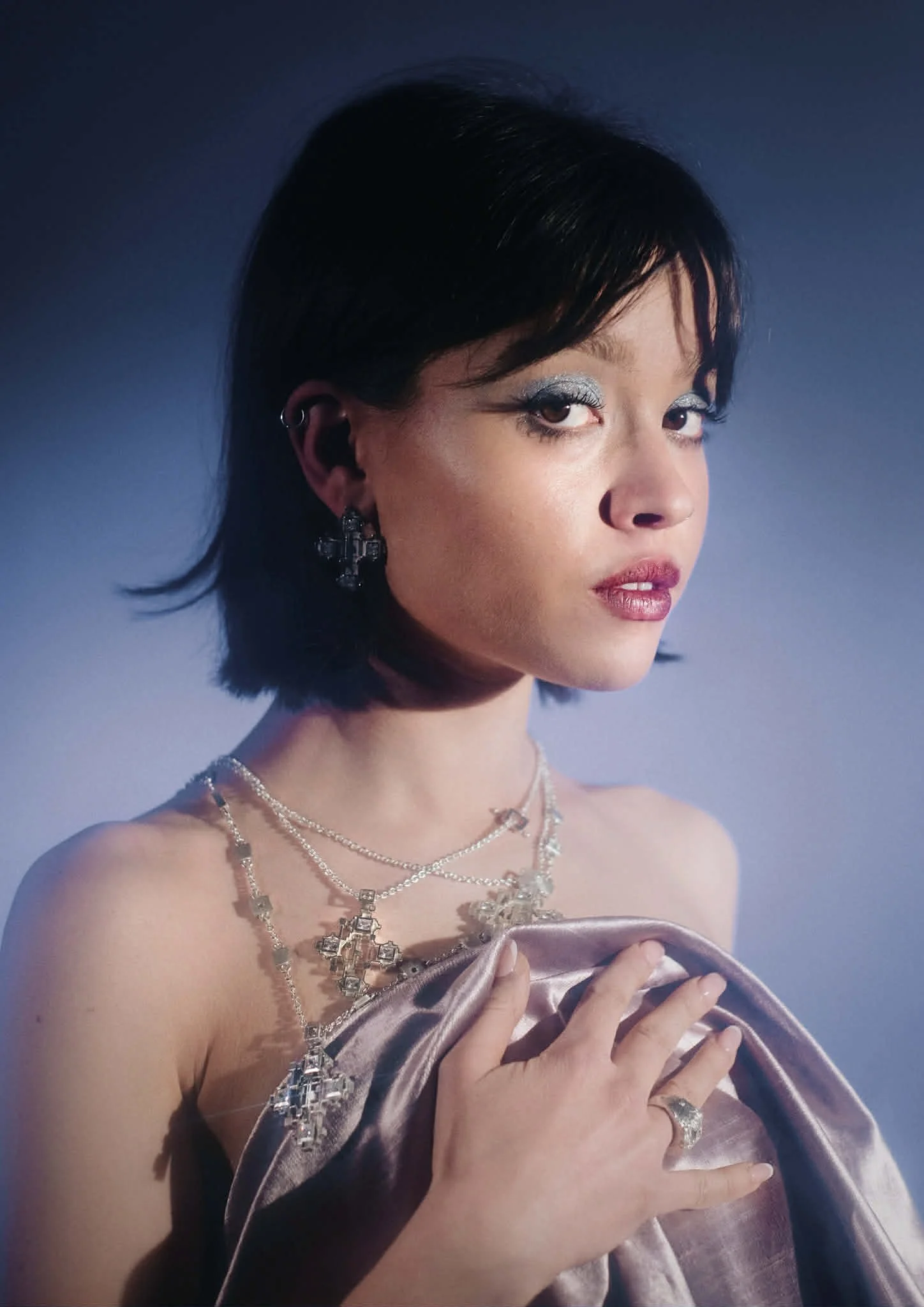 A woman with short black hair and silver makeup on her eyes, wearing multiple silver necklaces and earrings, holding a shiny satin fabric, against a plain gradient background.