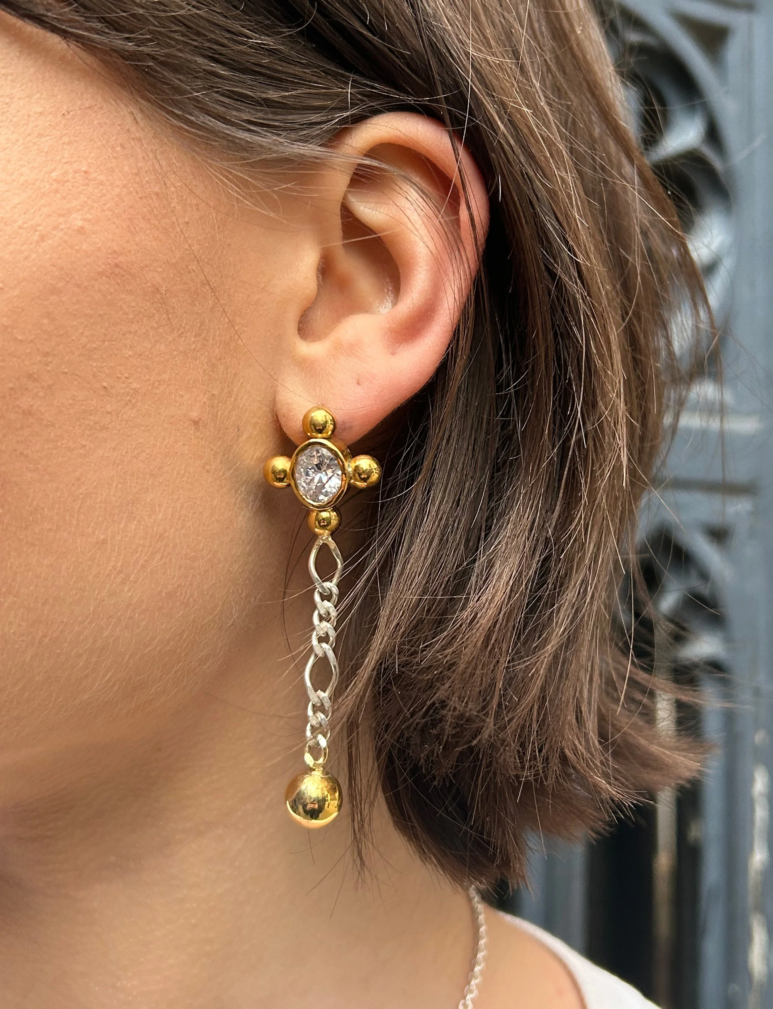 Close-up of a woman's ear with a gold and diamond earring, brown hair, and a necklace, against a blurred background.