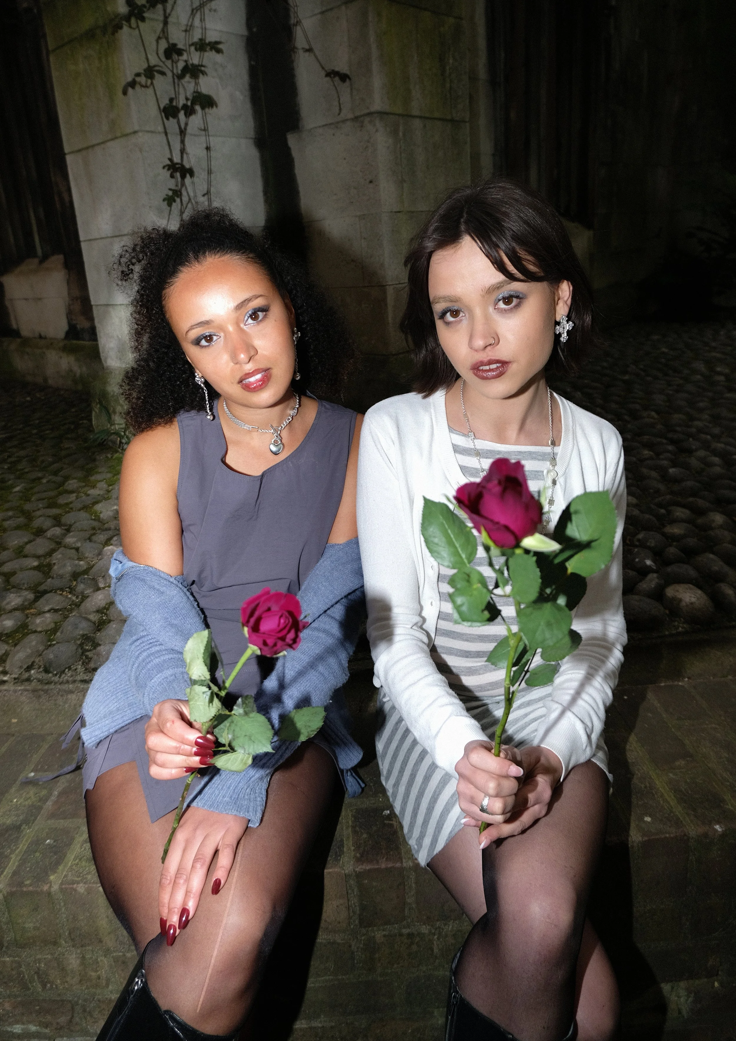 Two women sitting outdoors at night, holding roses, dressed in stylish clothing and jewelry, with a dark background and a stone wall.