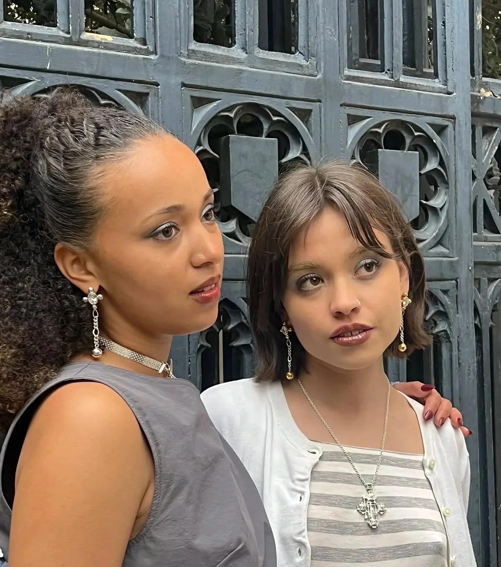 Close-up of two women standing close together outdoors in front of a decorative black metal gate. The woman on the left has curly dark hair, wearing a grey sleeveless top, and sparkling earrings. The woman on the right has straight dark hair, wearing