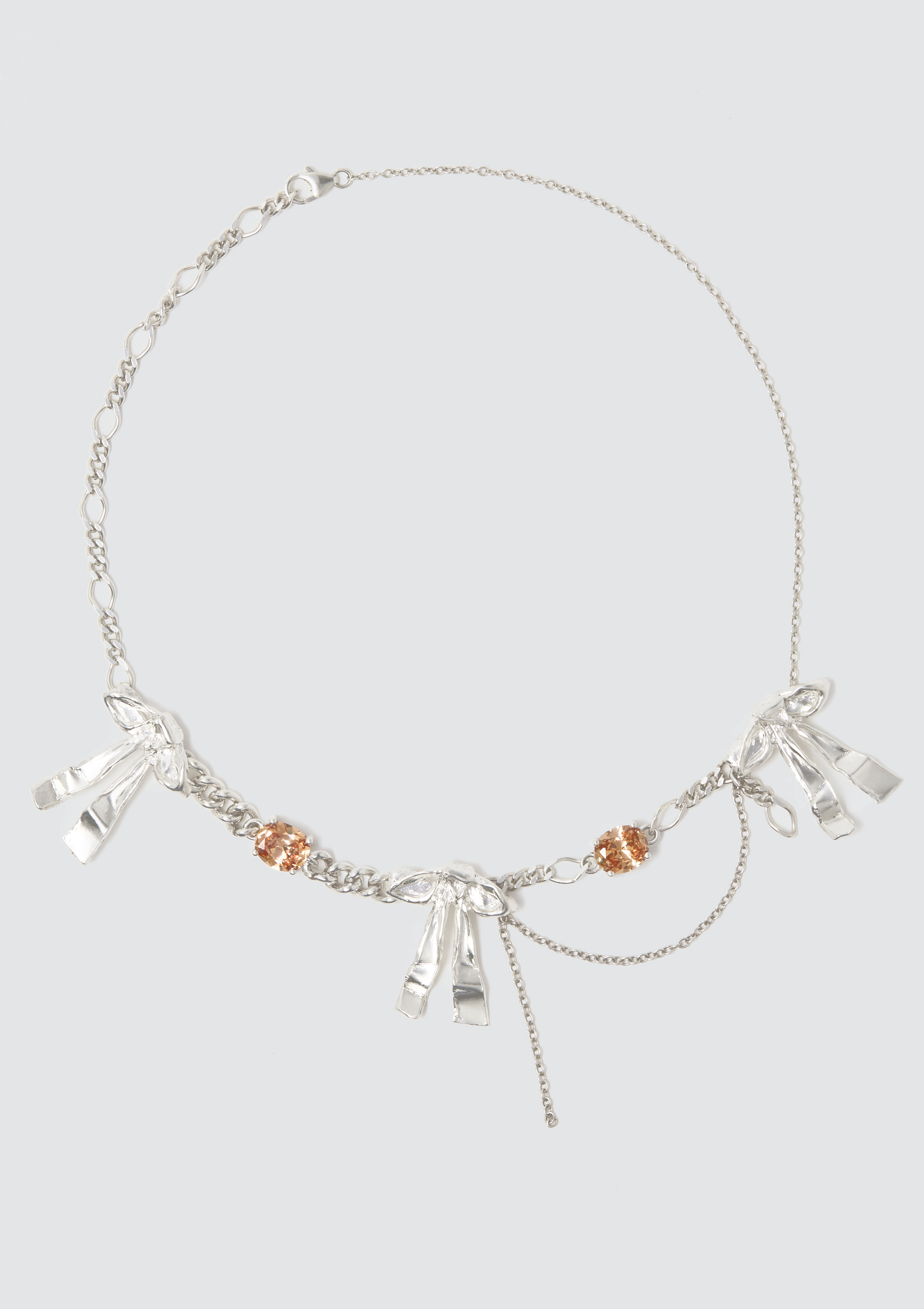 Close-up of a silver necklace with leaf and bow-shaped charms, accented with orange gemstones, on a light gray background.