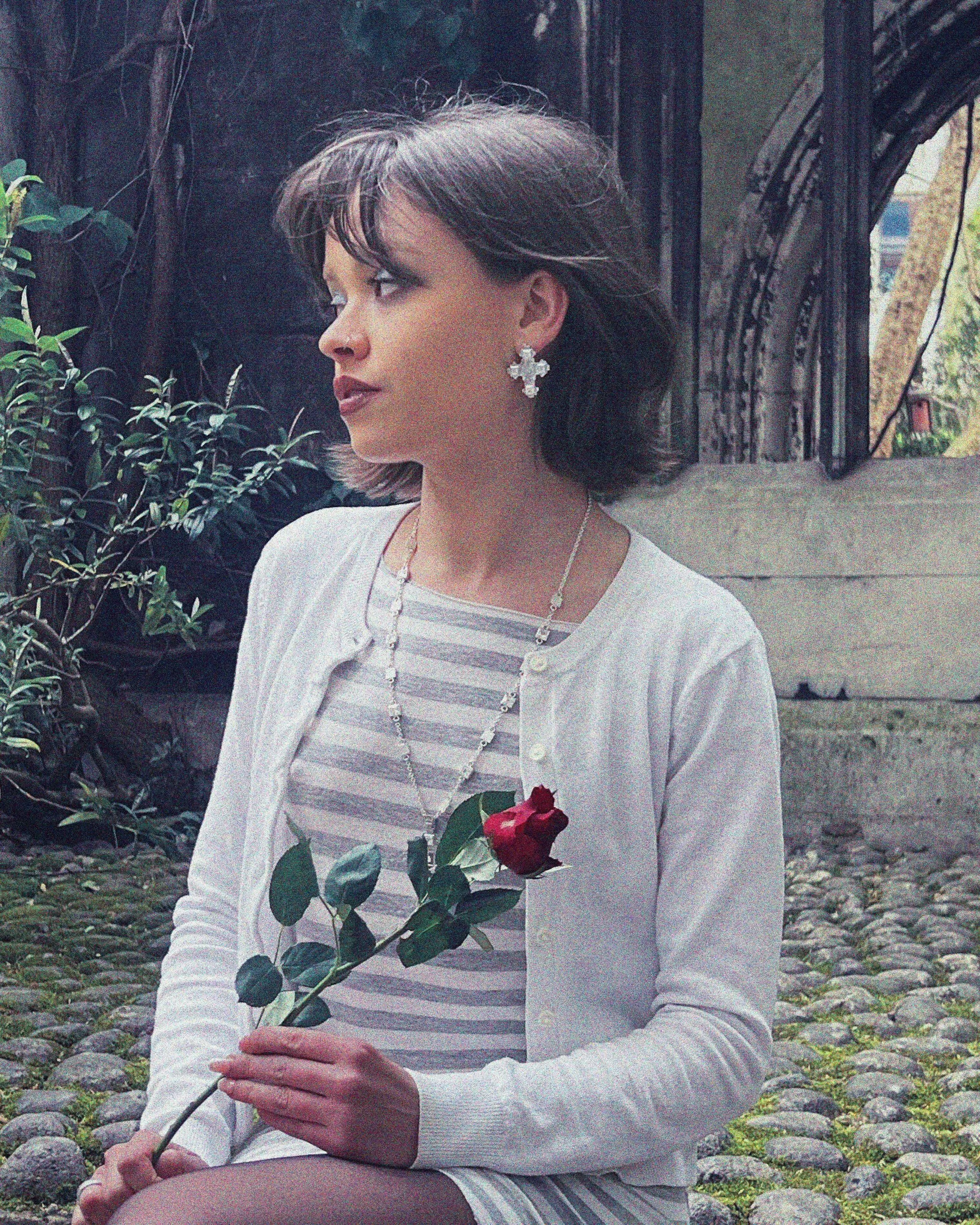 A woman sitting outdoors on a stone path, holding a red rose, with trees and foliage in the background. She is wearing a white cardigan over a striped top, and has earrings and jewelry visible.