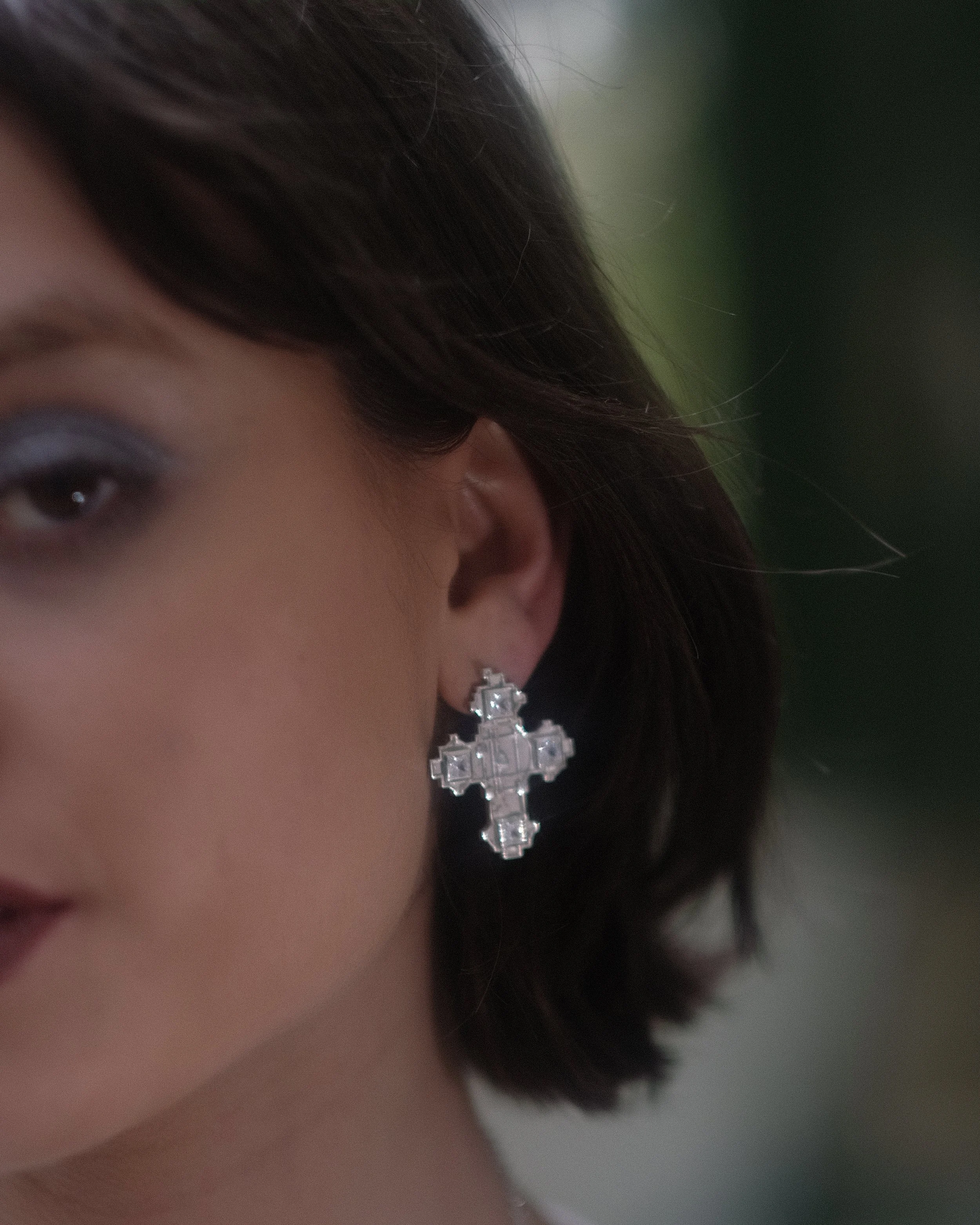 Close-up image of a woman wearing large cross-shaped earrings with square inlays, and part of her eye visible, with dark hair styled around her face.