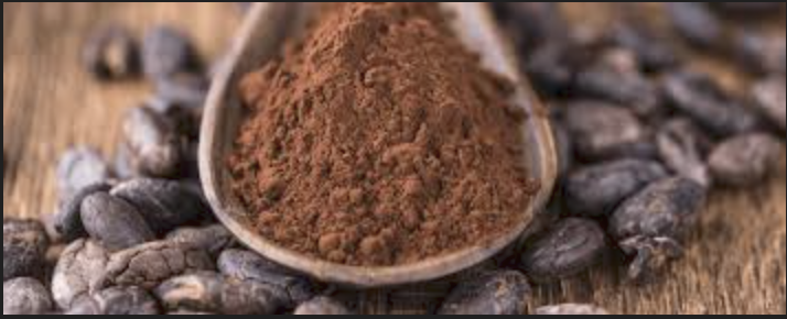 Cacao powder in a spoon surrounded by cacao beans on a wooden surface.