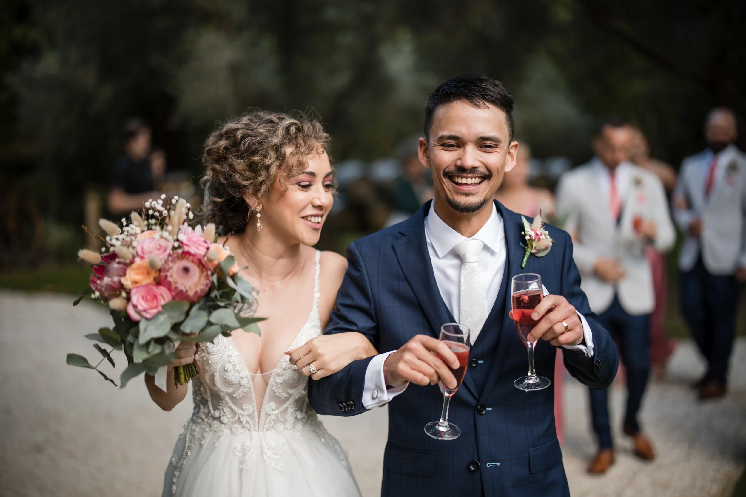 A bride and groom smiling and holding glasses of champagne at their wedding reception outdoors, with guests in the background.