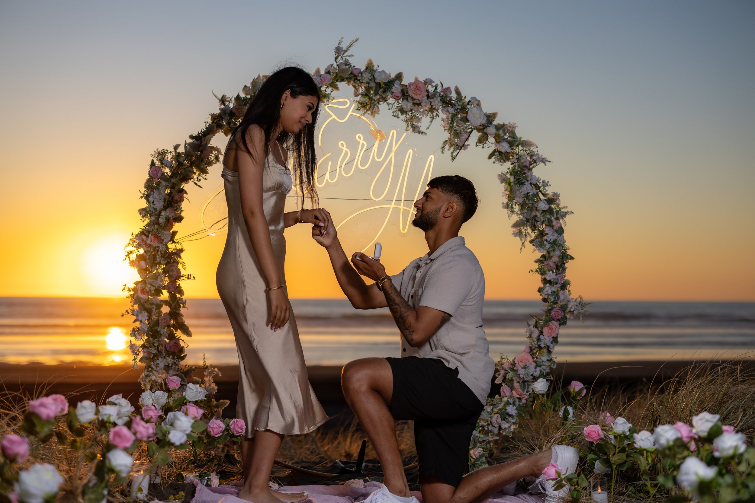 A couple proposes at a beach during sunset. The man is on one knee holding a ring, while the woman stands in front of him. They are under a floral arch with a neon sign behind them that reads 'marry me.' Flowers are arranged around the arch, and the scene is illuminated by the setting sun with ocean waves in the background.