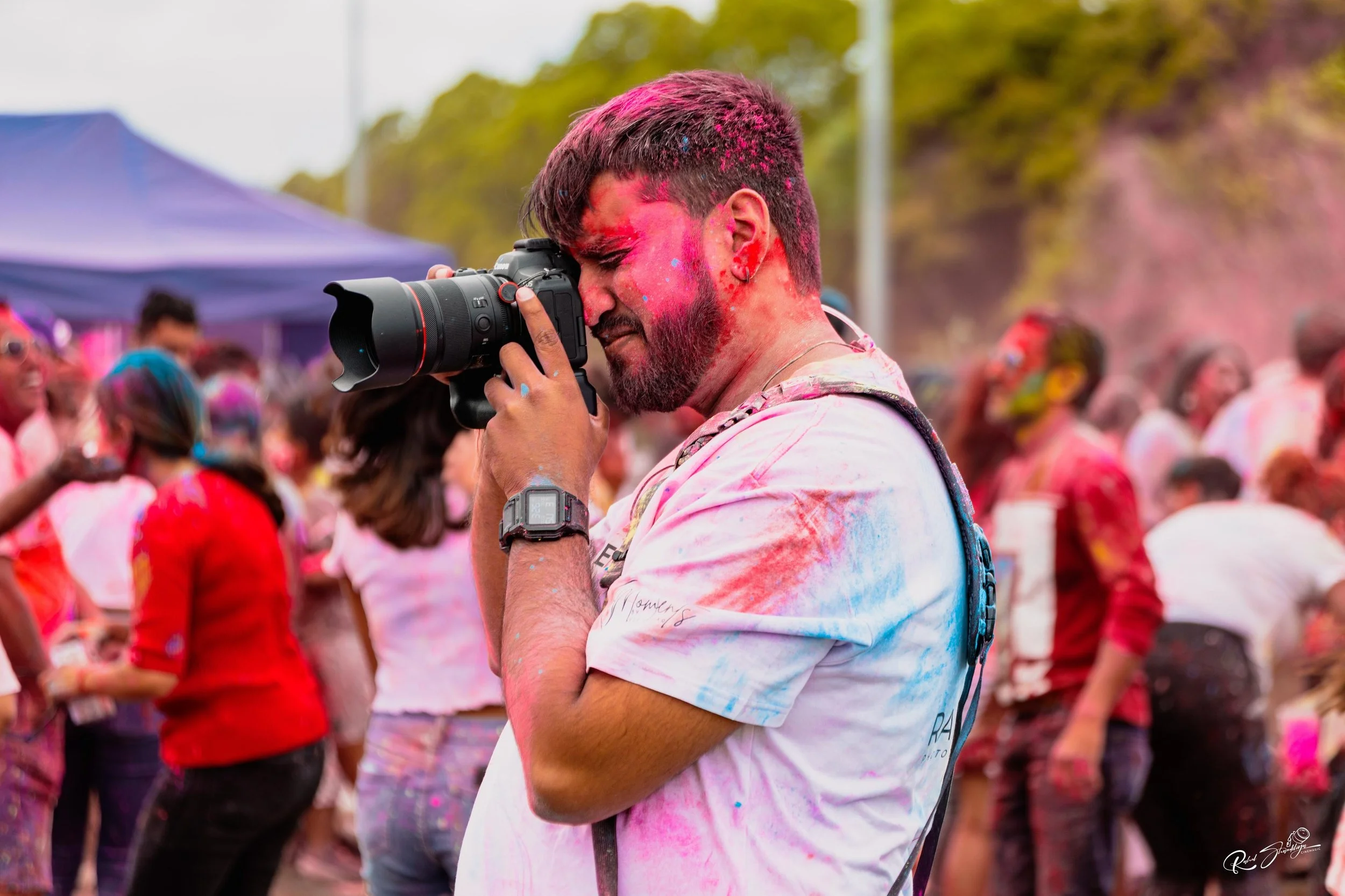 A man using a camera during a color festival, with pink, red, and blue powders covering his face and shirt, surrounded by people also covered in colorful powders.
