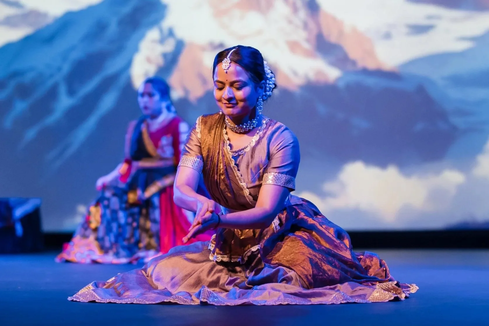 Women in traditional Indian attire performing a dance on stage with a mountain backdrop.