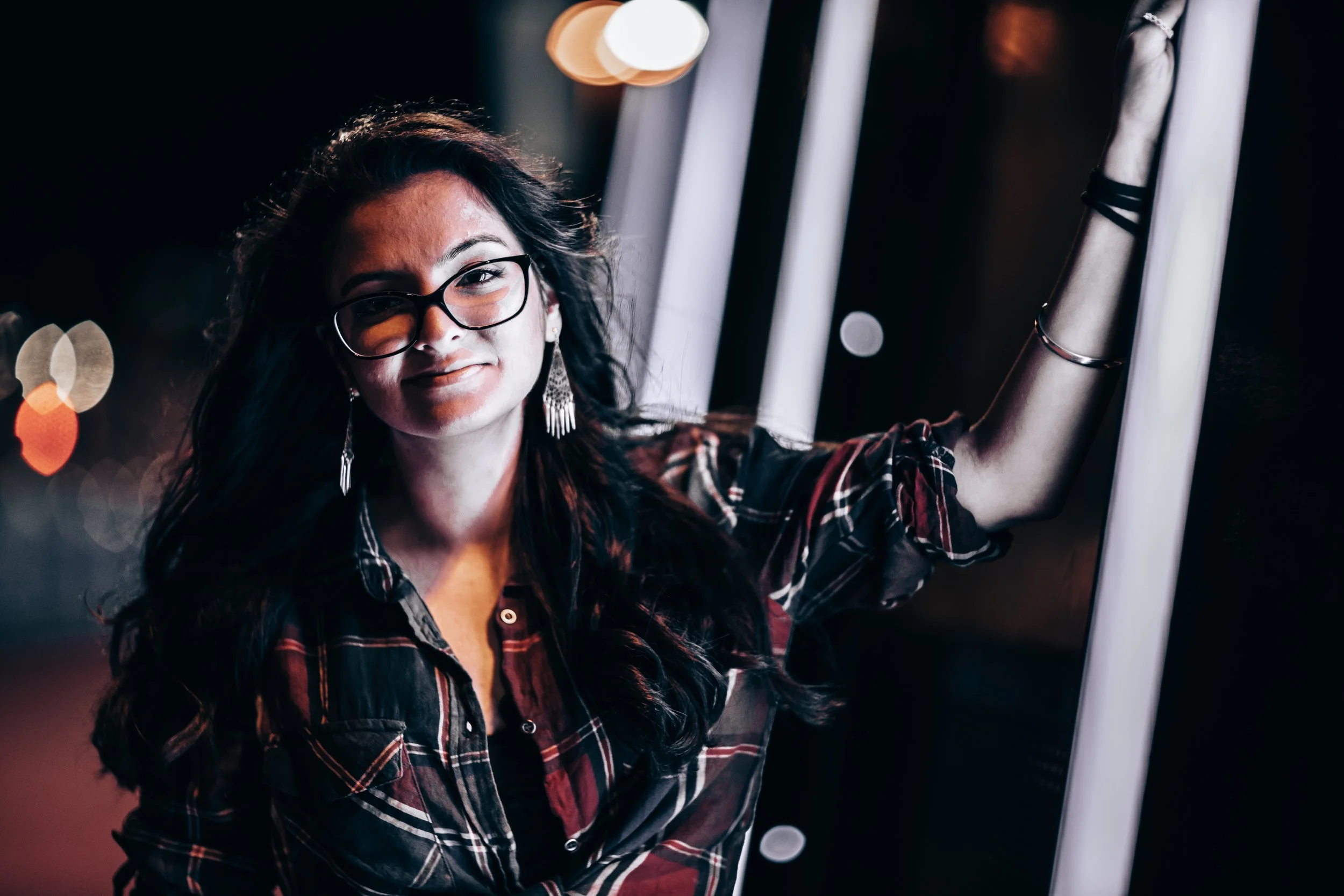 A woman with long dark hair, glasses, and earrings smiling and touching a white illuminated structure in a dark setting with bokeh lights in the background.
