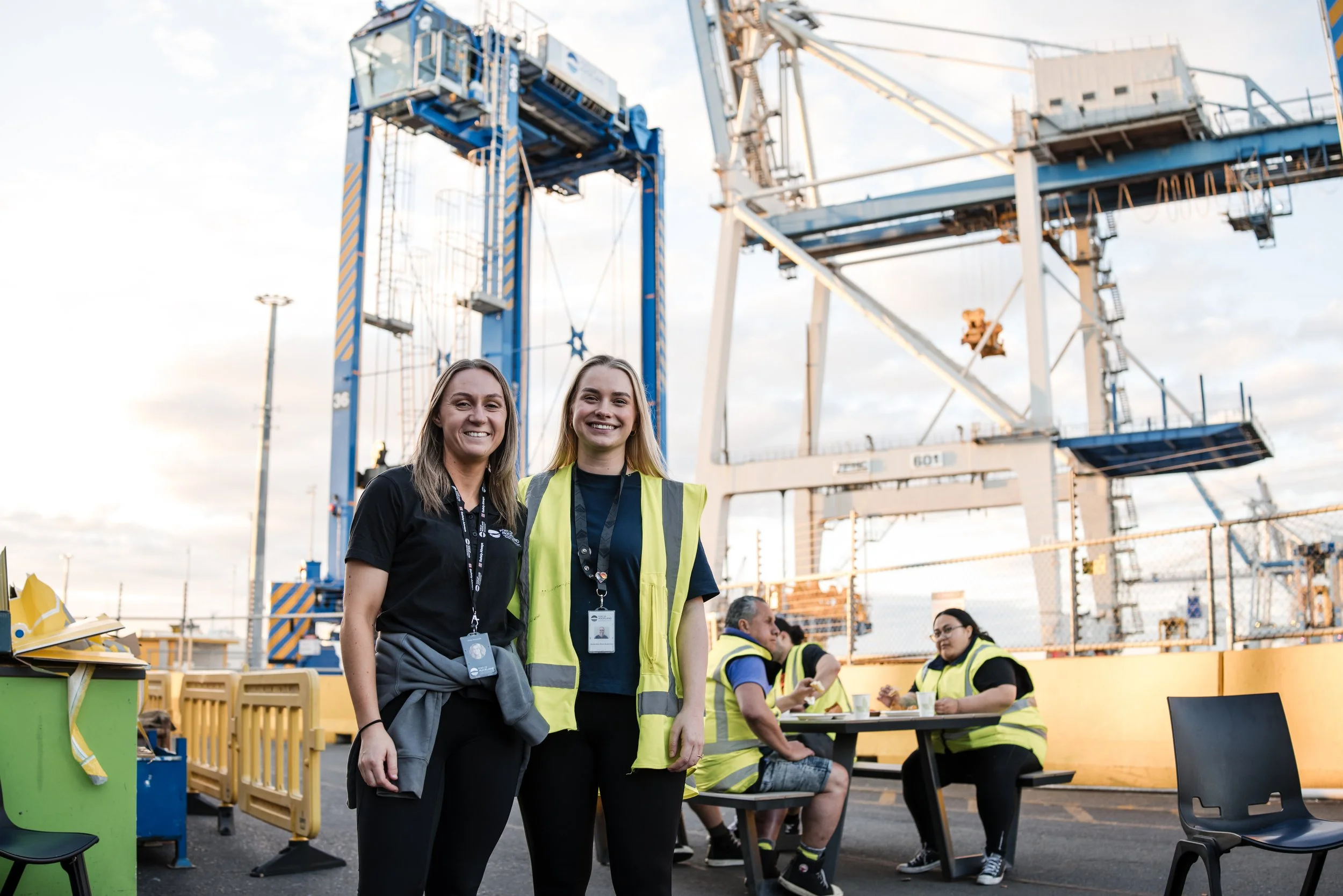 Two women smiling at the camera at an outdoor event with amusement park rides in the background. One woman is wearing a black shirt with a lanyard, and the other is wearing a yellow safety vest. Several other people are visible, some wearing yellow safety vests, sitting or standing around tables.