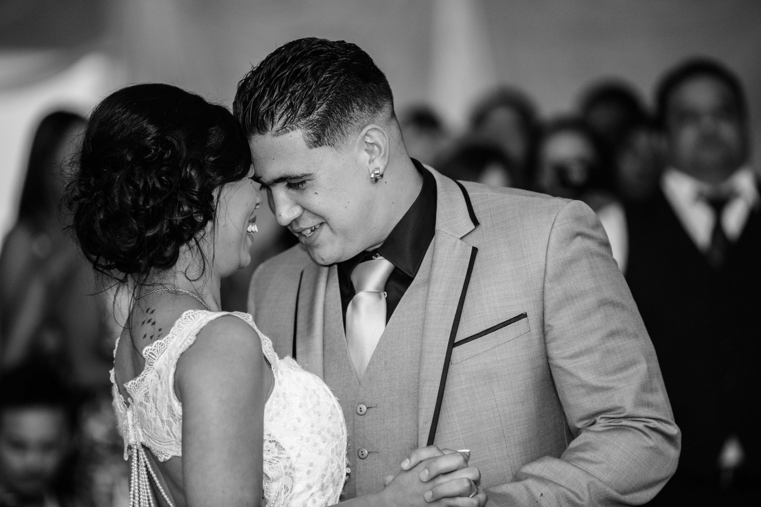 A black-and-white photo of a couple dancing closely at a formal event, with the woman's face partially obscured, wearing a white lace dress, and the man in a suit and tie, with his face touched to hers.