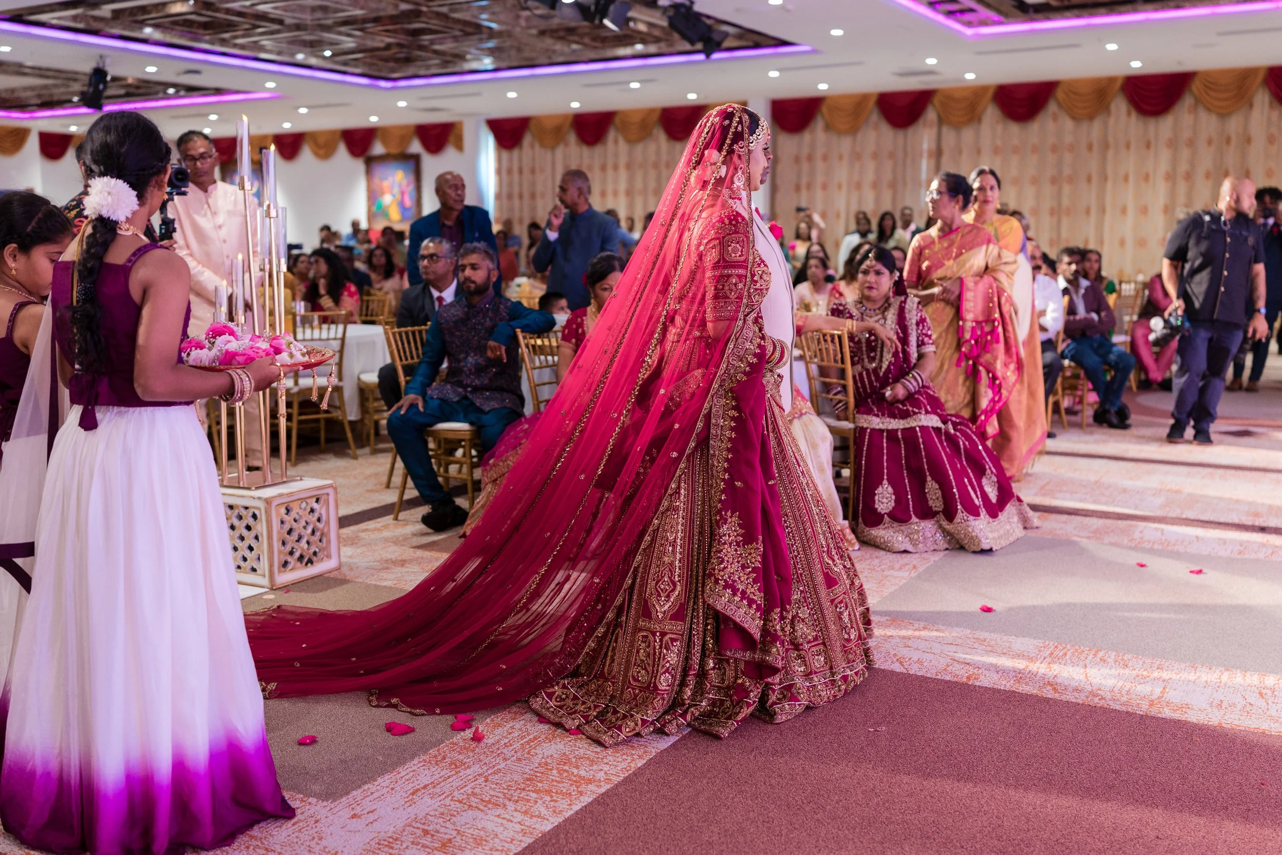 A bride in a red and gold traditional wedding dress with an elaborate embroidered veil walking in a decorated wedding hall, with seated guests and women in colorful sarees observing the ceremony.