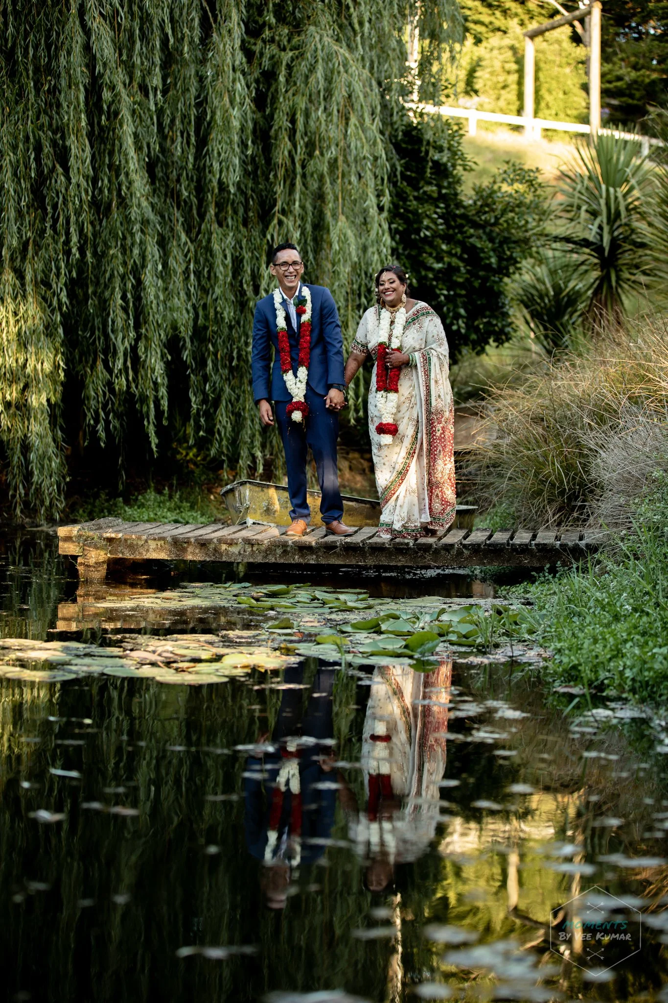 A couple in wedding attire walking hand in hand on a wooden bridge over a pond with lily pads, surrounded by lush greenery and tall trees, smiling and enjoying their moment.