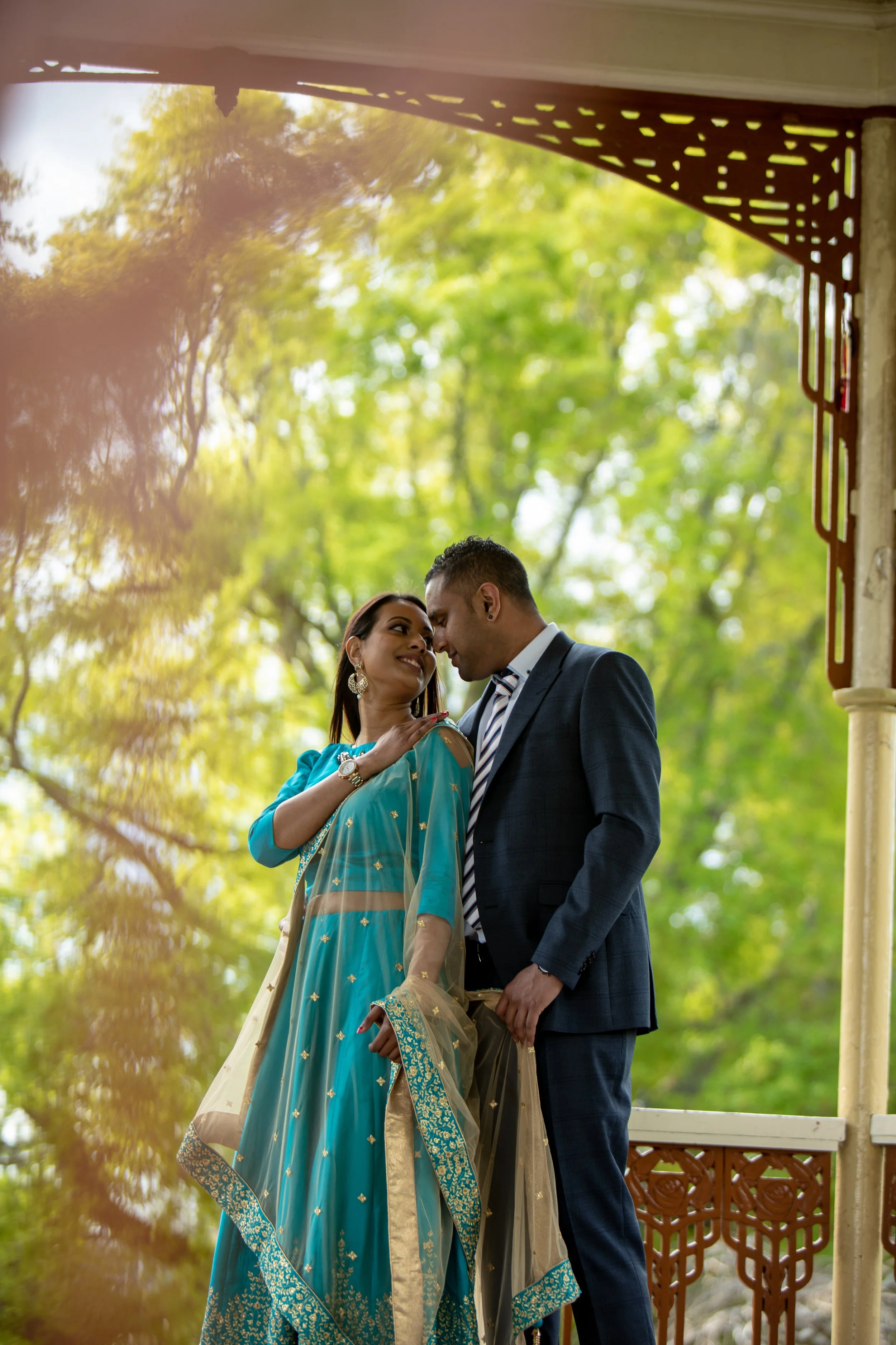 A couple dressed in formal attire, standing close and touching foreheads, under a gazebo with green trees and bright sunlight in the background.