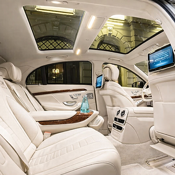 Interior of a luxury car with beige leather seats, a glass of water on the center armrest, and a panoramic sunroof, with screens on the back of the front seats.