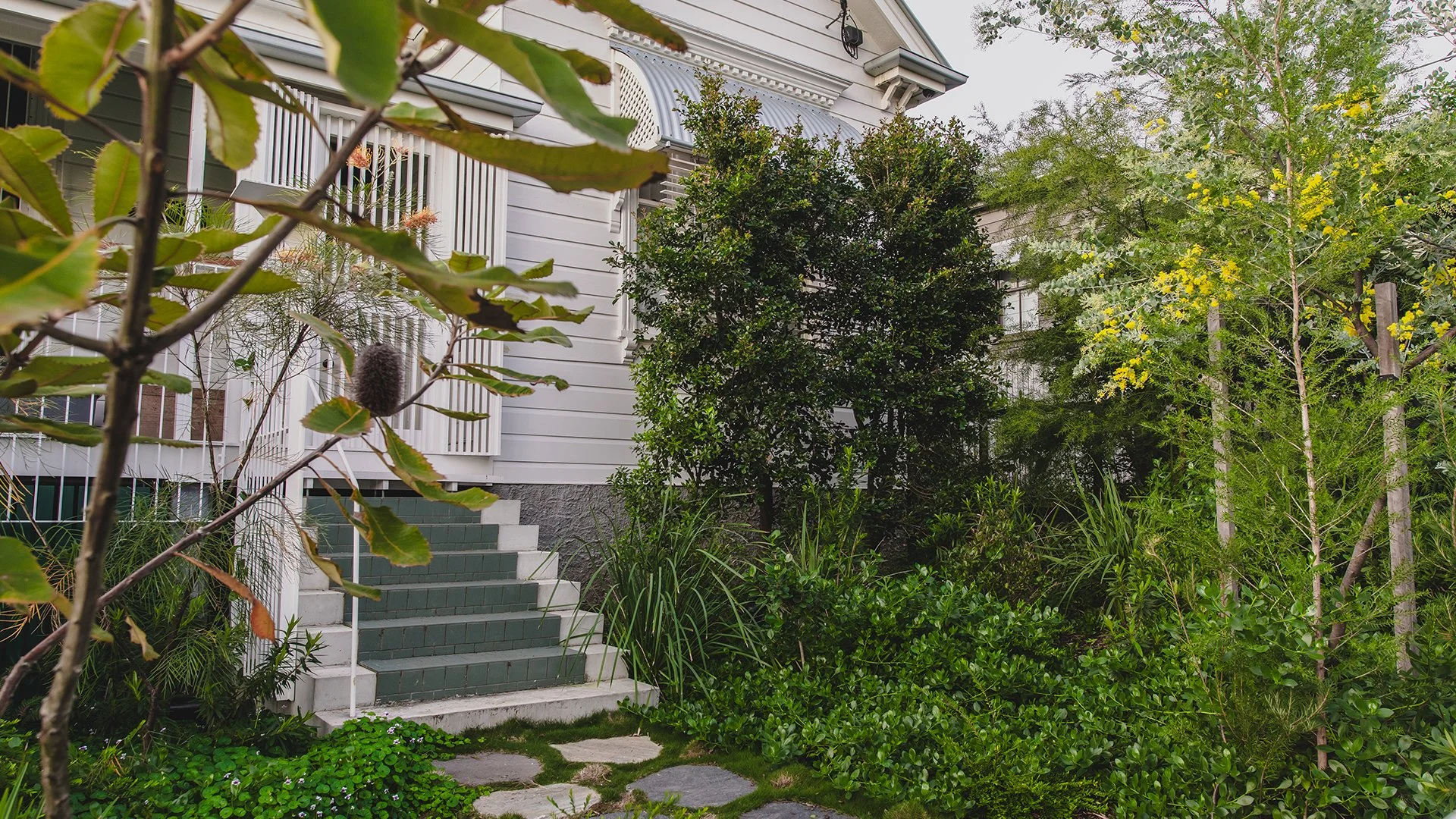 A house with white siding and green stairs leading to the entrance, surrounded by lush greenery and trees.