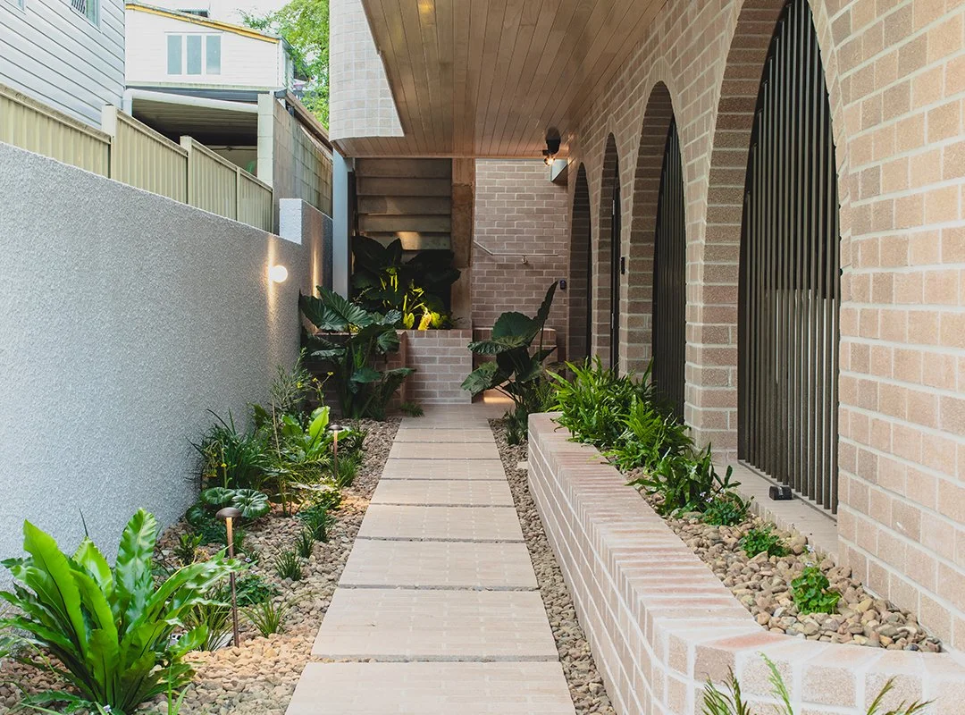Narrow outdoor walkway on the side of a brick house, with plants and garden lights along the path, leading to stairs at the back.