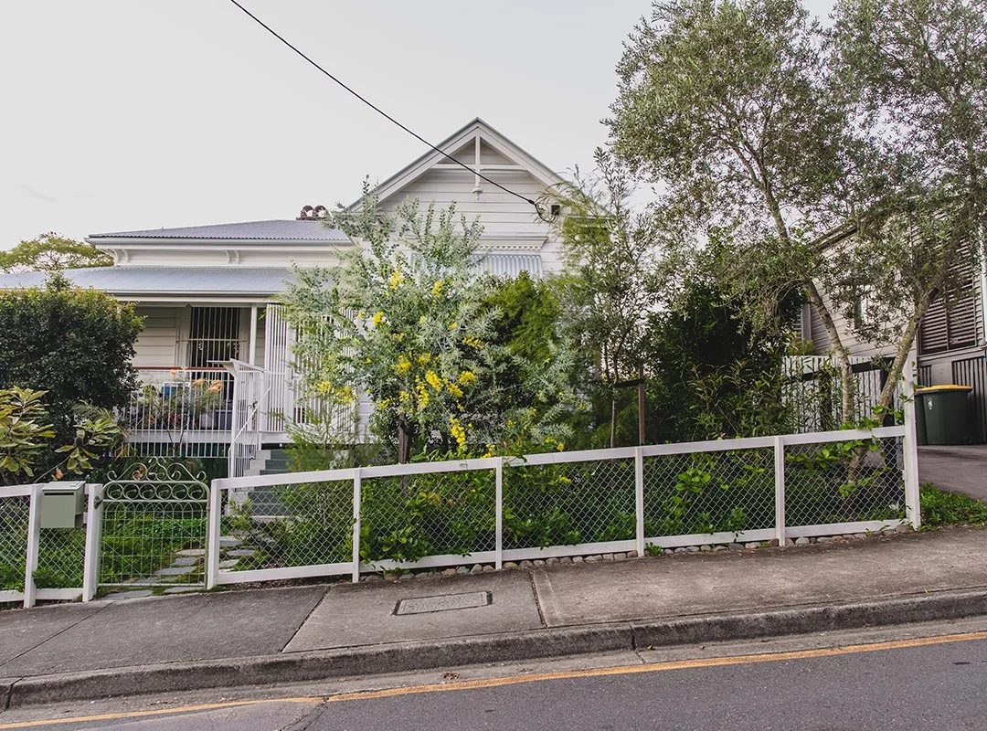 A white house behind a white metal fence with a sidewalk in front, surrounded by trees and greenery.