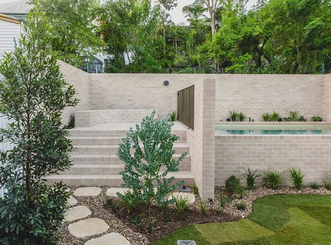 Backyard with brick steps leading up to a patio area, surrounded by green plants and trees, with a raised pool or hot tub to the right, and a well-manicured lawn in the foreground.