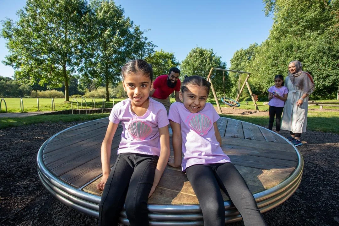 Two young girls in light purple shirts sitting on a round wooden playground equipment with adults and children in the background, outdoors in a park with trees and a swing set.