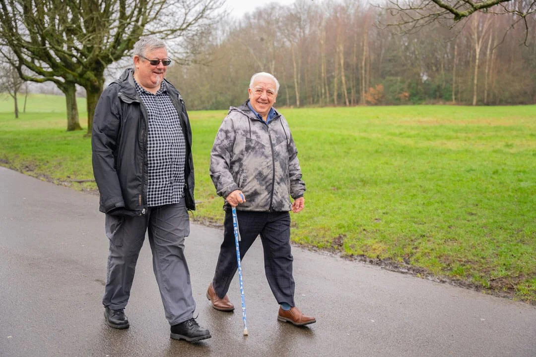 Two elderly men walking on a paved path in a park during daytime, one using a cane, the other smiling and wearing sunglasses, with trees and grass in the background.