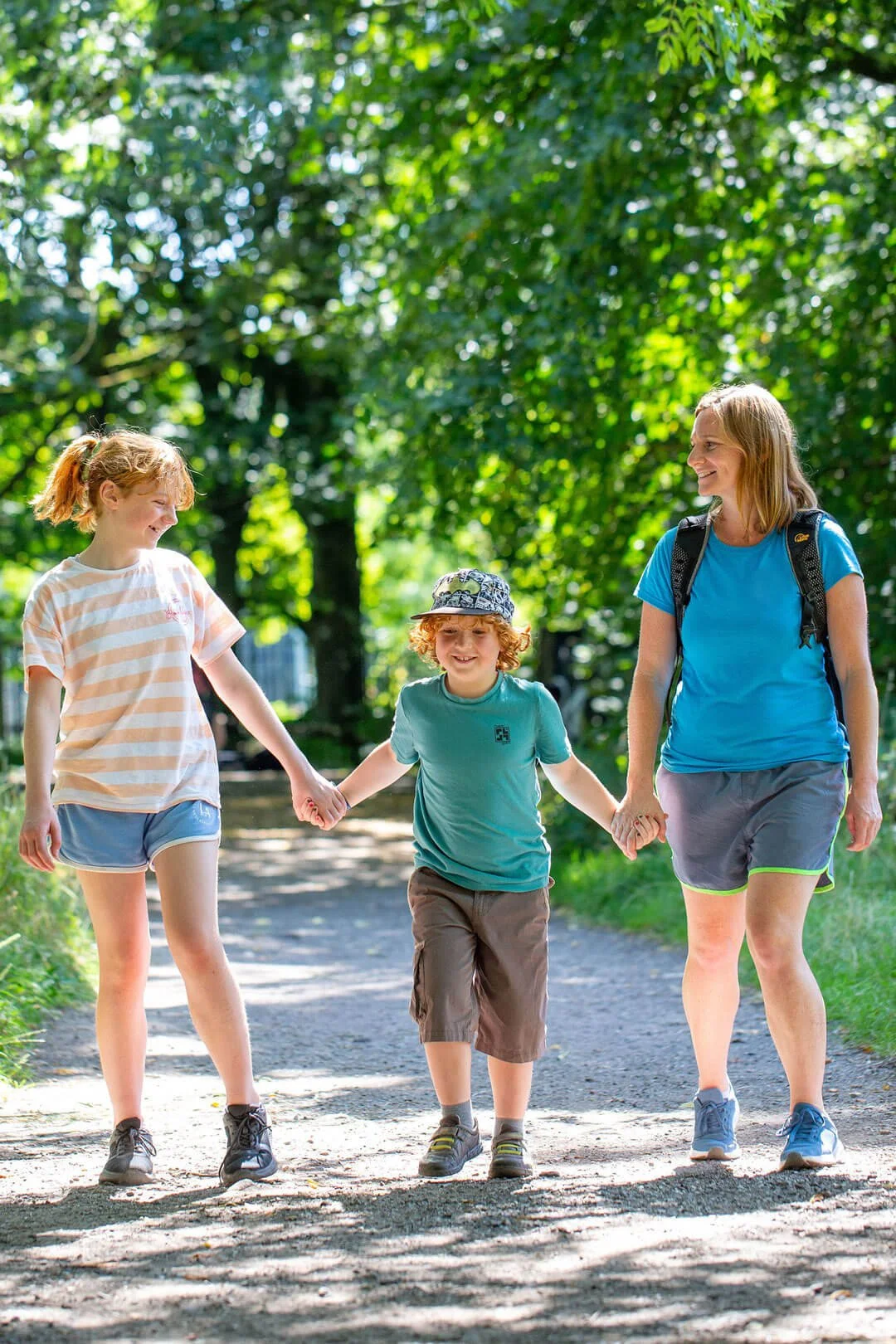 A woman and two children holding hands and walking on a forest trail, enjoying a sunny day outdoors.