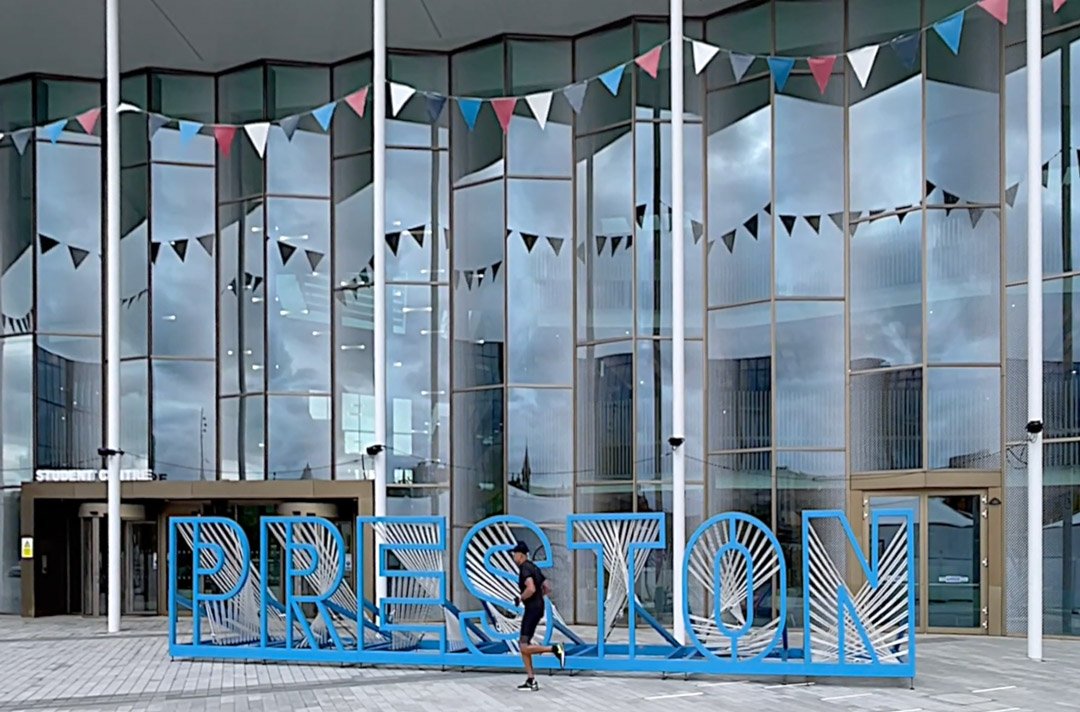 Large blue 3D letters spell out "PRESTON" in front of a modern glass building with triangular pennant banners hanging above.