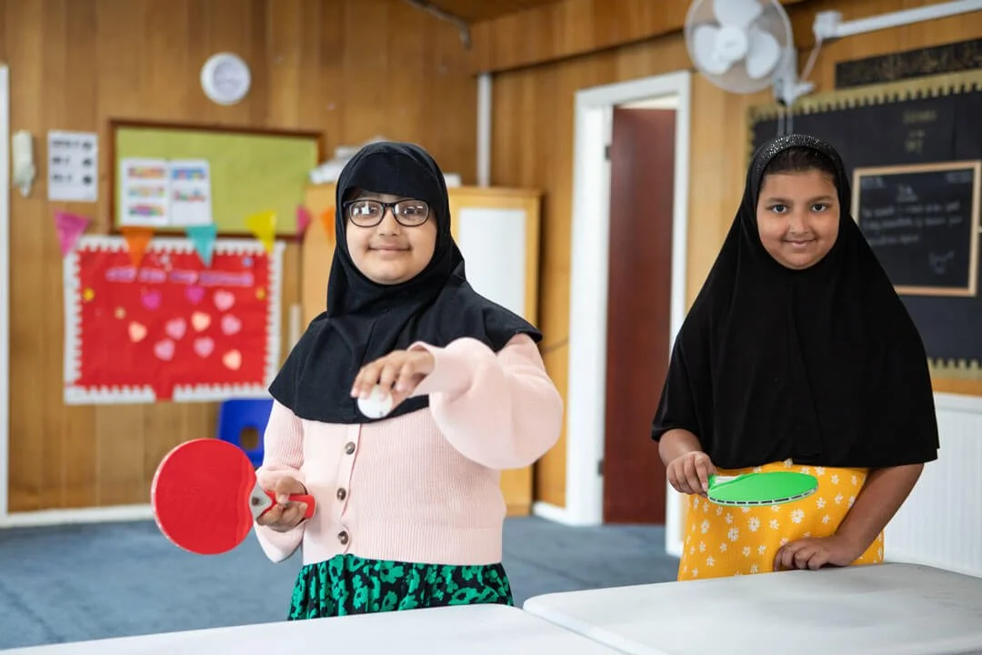 Two young girls wearing hijabs playing table tennis in a classroom.