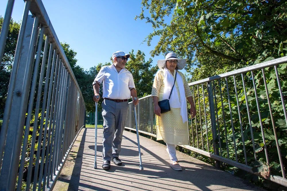 An elderly man and woman walk on a sunny outdoor pathway, with the man using crutches and the woman wearing a wide-brimmed hat.