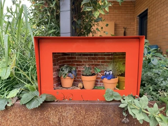 A red miniature greenhouse with potted plants and a blue fuzzy creature inside, surrounded by green plants and garden elements.